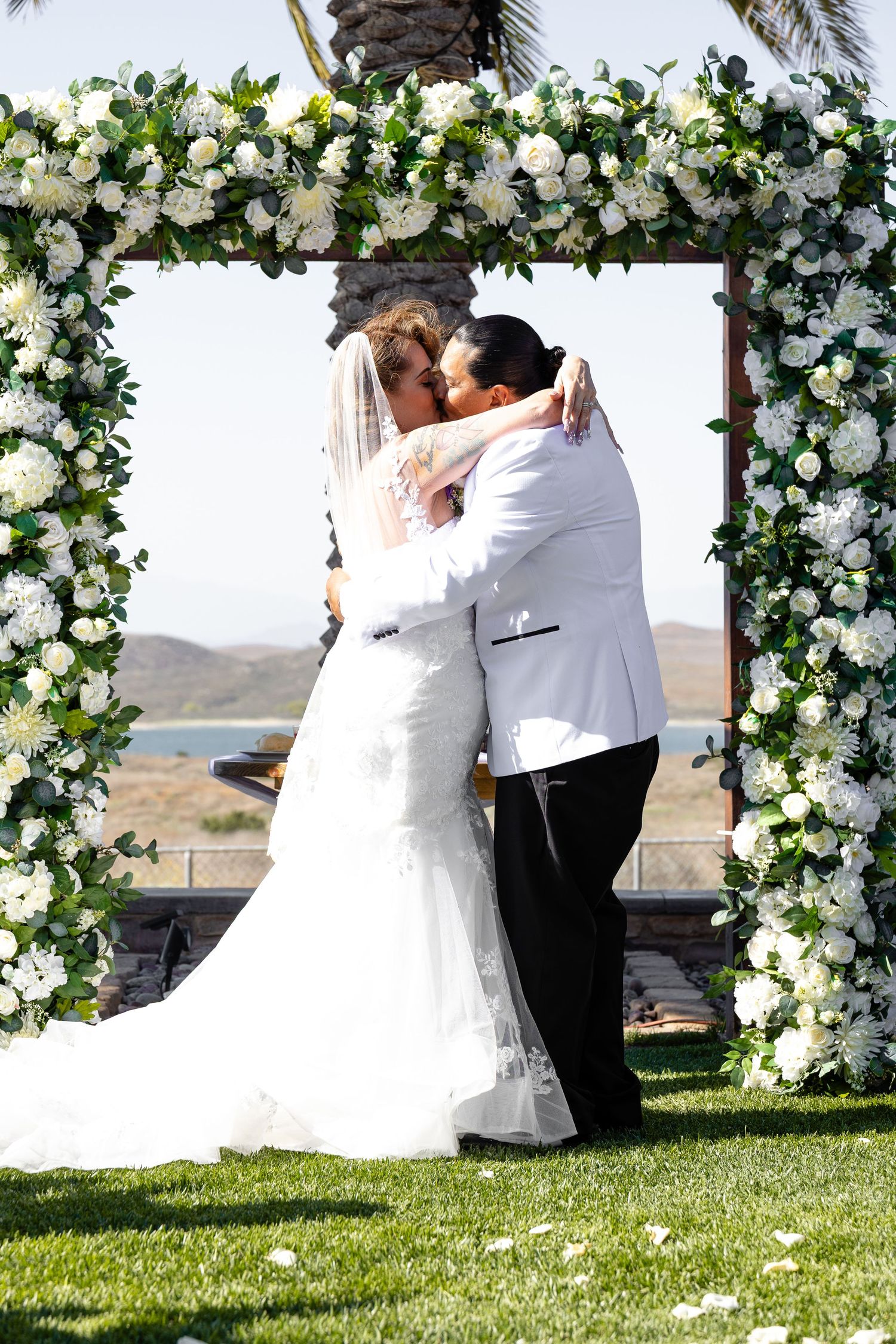 A romantic wedding ceremony under a beautiful floral arch decorated with white roses and greenery on a sunny day.