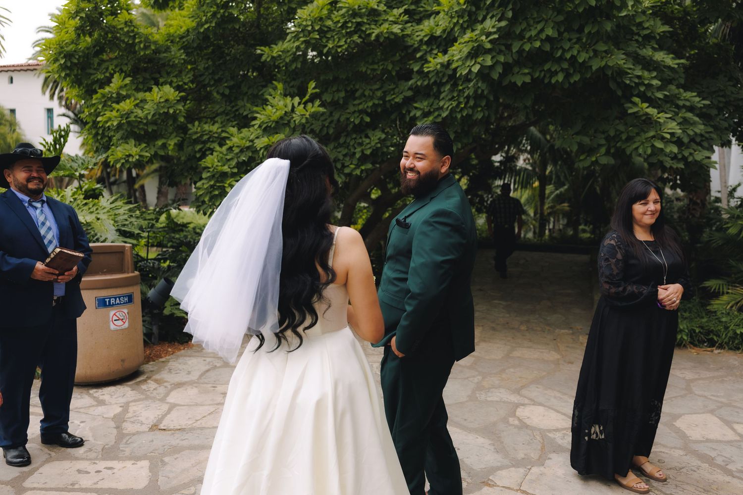 A wedding celebration outdoors with guests gathered on a stone patio surrounded by green trees.