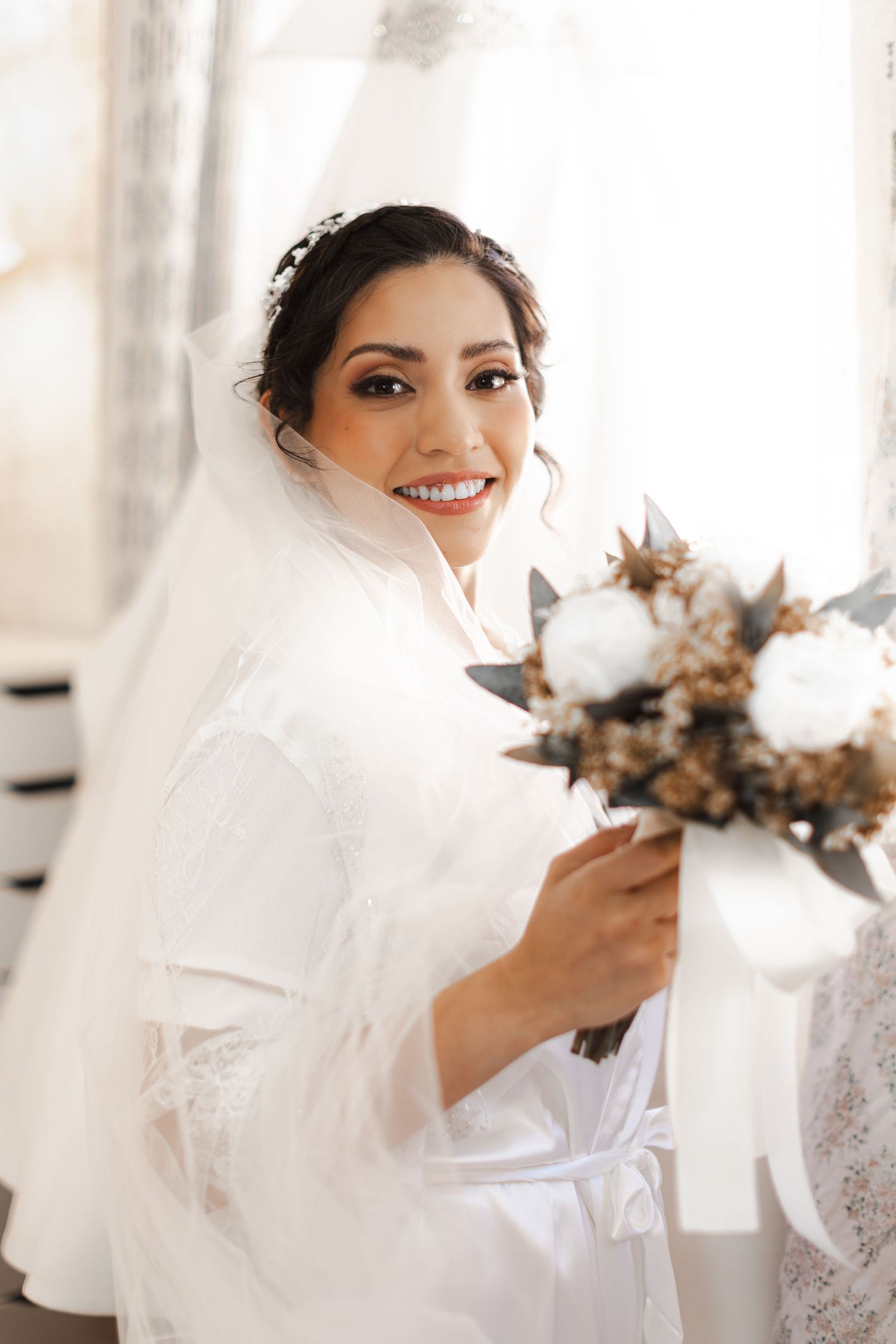A bride in a white wedding gown and veil holds a dried floral bouquet while smiling in a bright indoor setting.