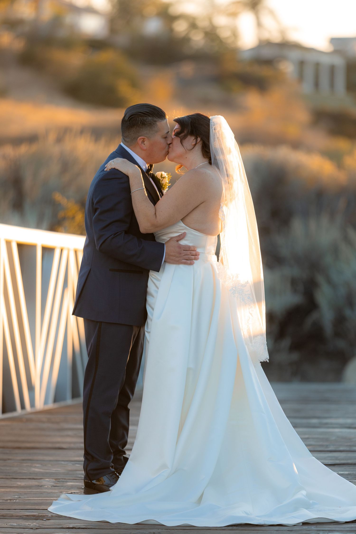 Romantic wedding couple sharing a tender kiss on a wooden boardwalk at sunset.