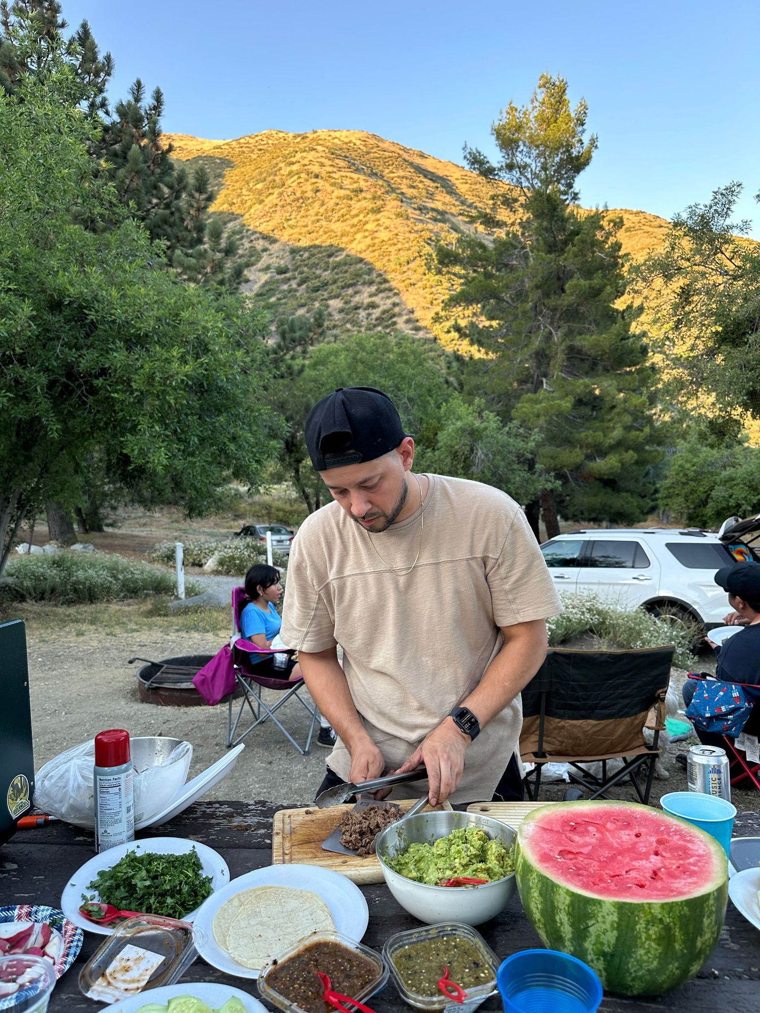 A picnic scene in front of mountains shows food prep at a table with watermelon, salads and other dishes during sunset.