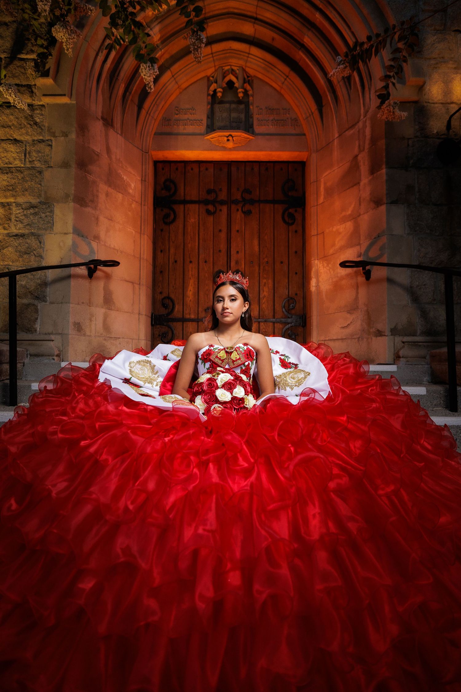 A stunning red quinceañera gown with a dramatic layered tulle skirt photographed in front of an ornate stone archway.