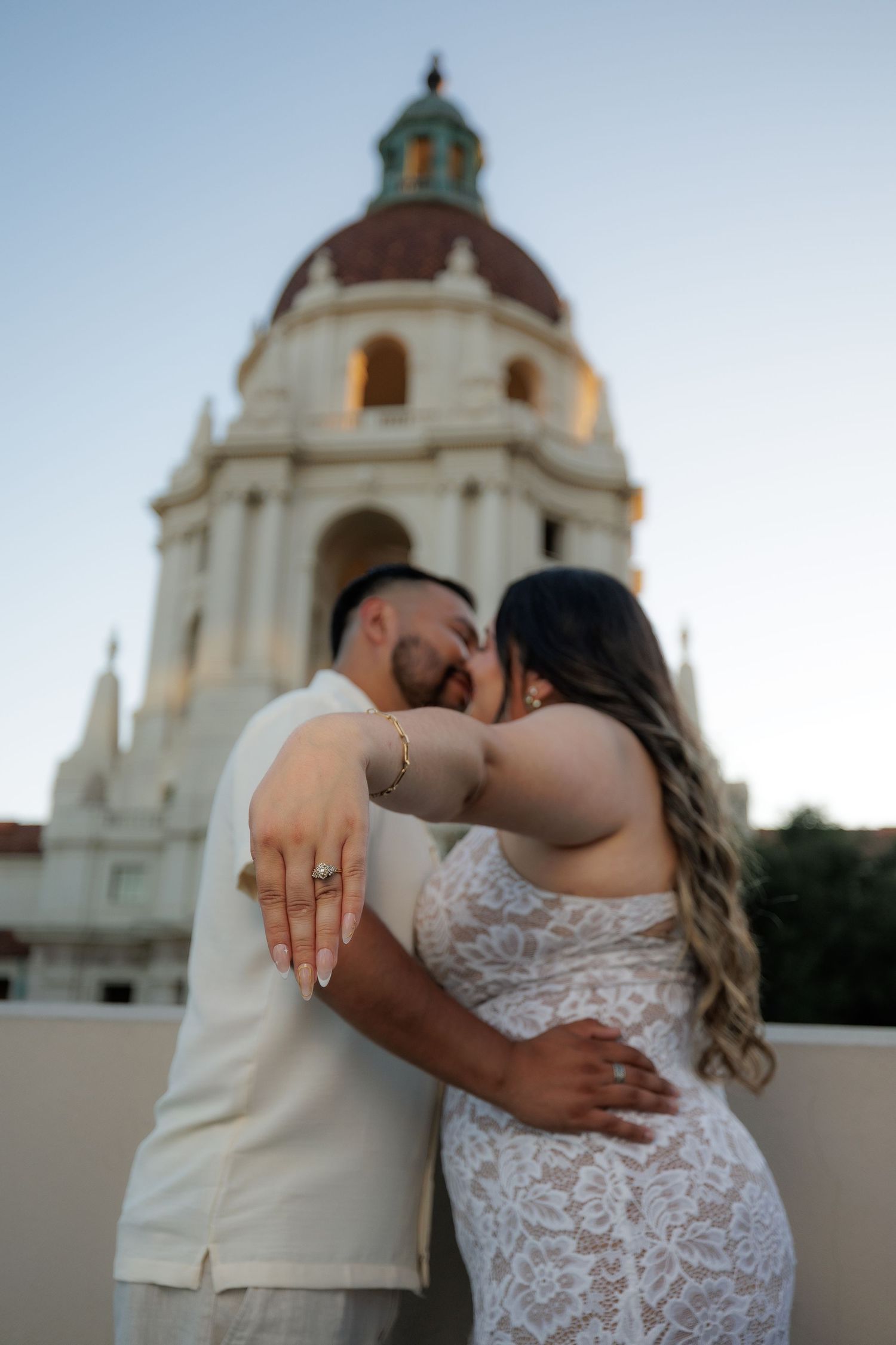A romantic couple in wedding attire shares an intimate kiss in front of a domed church building at sunset.