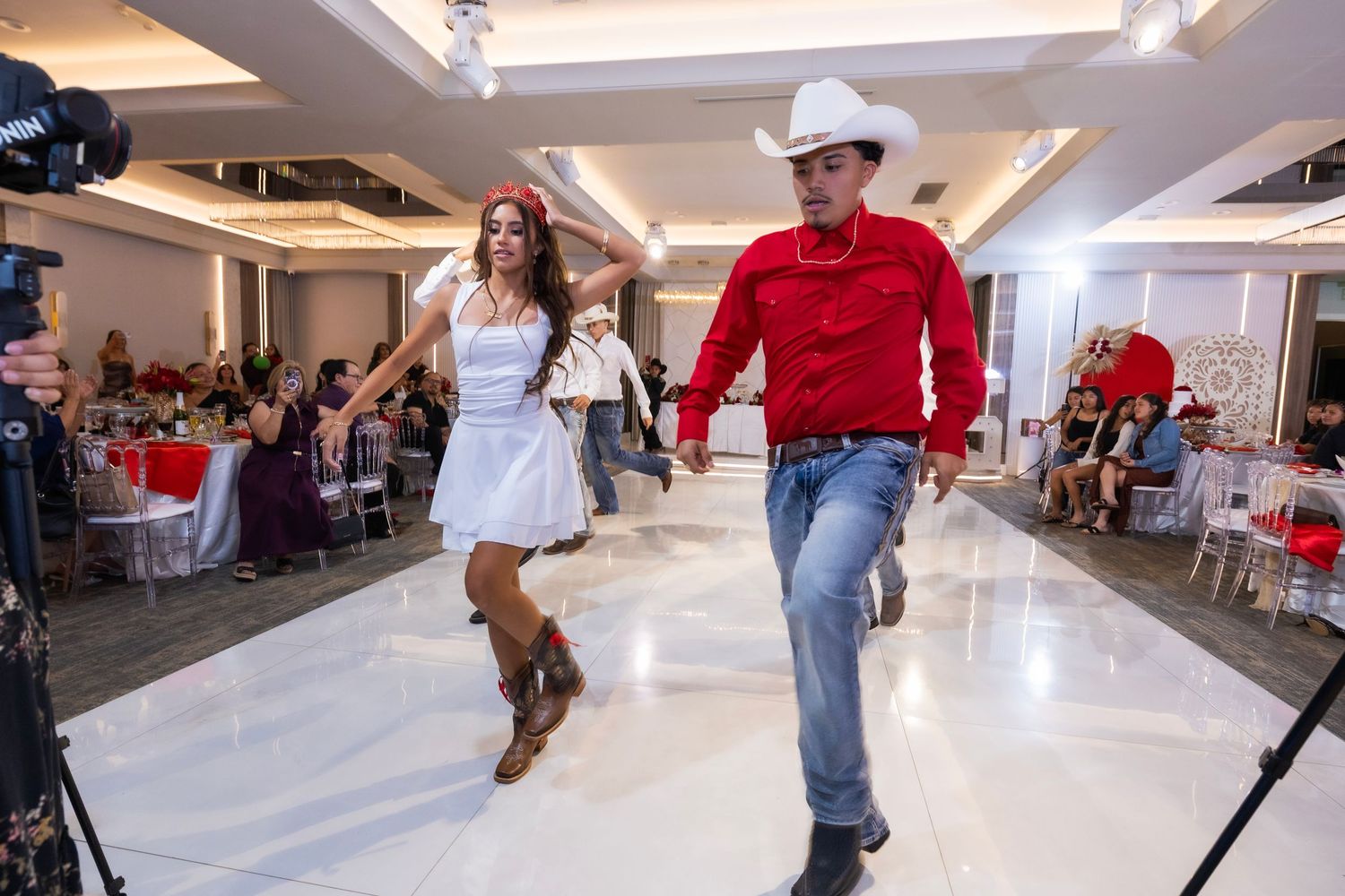 Dancers perform a lively country western dance on a shiny floor at an indoor event venue with tables in the background.
