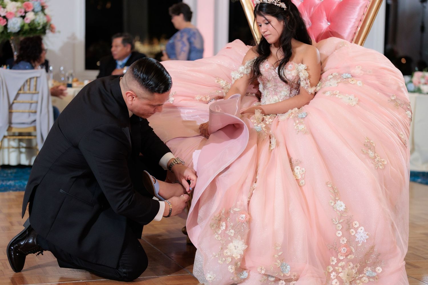 Dancers in pink gowns perform choreographed movements on a polished ballroom floor.
