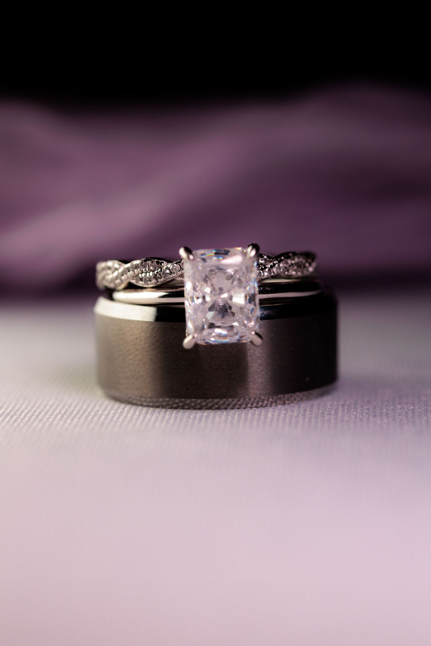 Two wedding rings rest on a textured purple surface with one featuring a princess cut diamond solitaire.