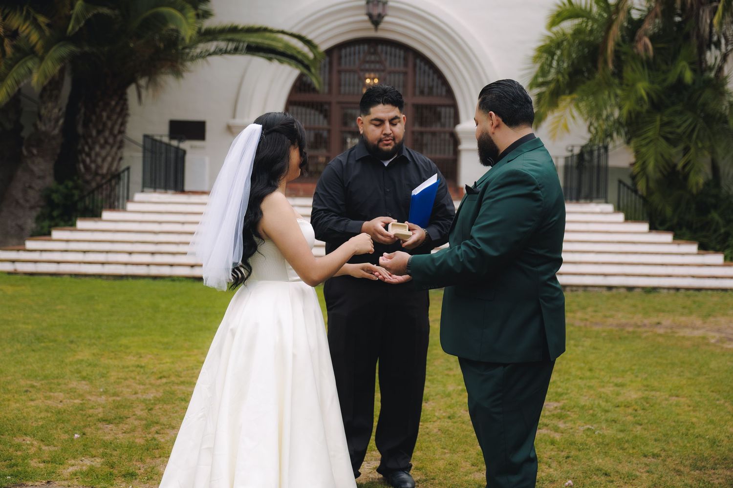 A wedding ceremony takes place outdoors with a couple and officiant standing in front of a white building.