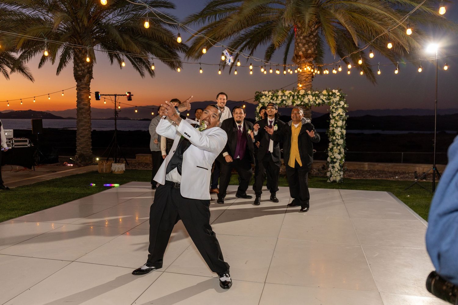 Wedding guests dancing and celebrating on an outdoor dance floor at sunset with palm trees and string lights overhead.