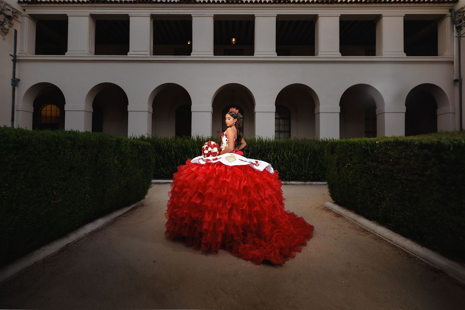 A dramatic red quinceañera dress creates striking visual contrast against a classical architecture backdrop in the garden.