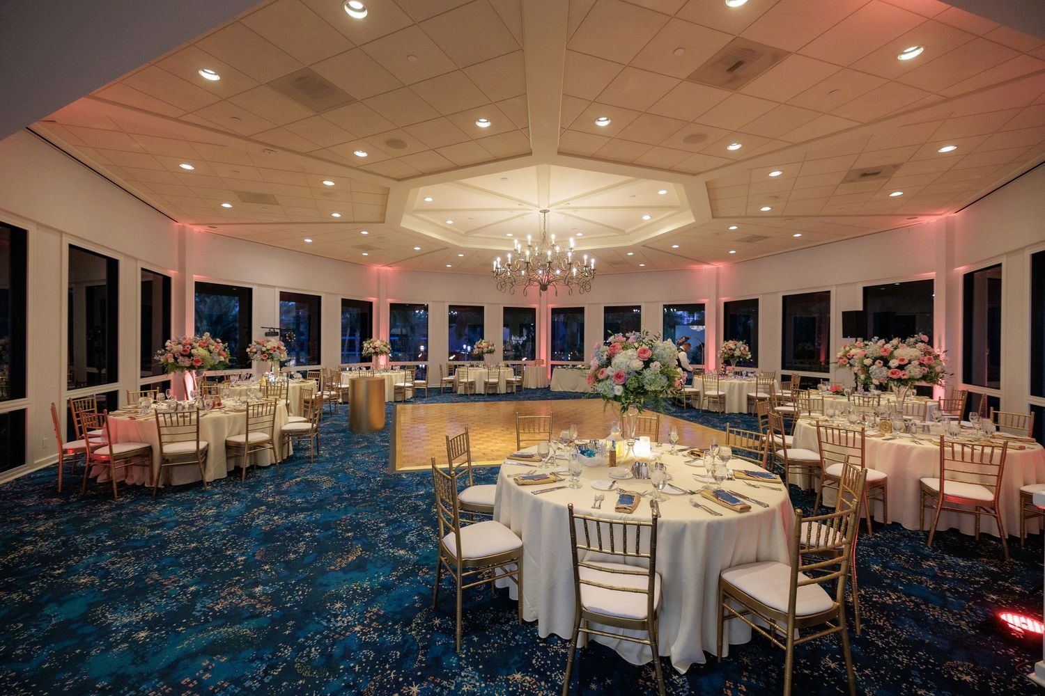 Elegant ballroom with crystal chandelier, round tables, and blue carpet set for a formal event.