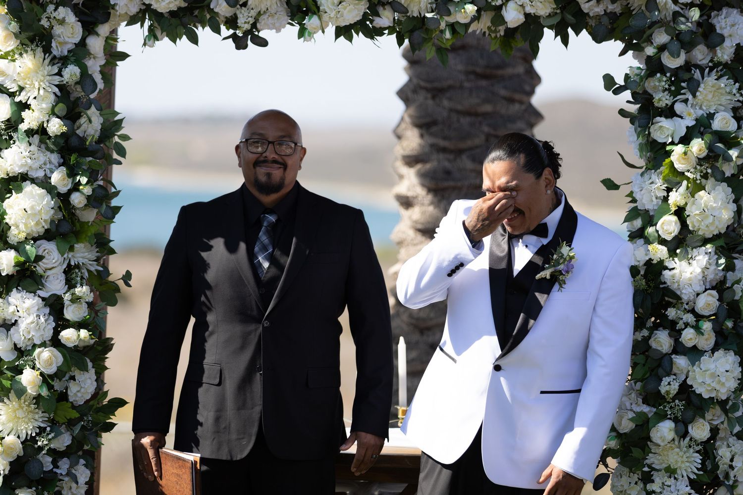 A group celebrates at an outdoor wedding ceremony under a white floral arch decorated with roses and greenery.