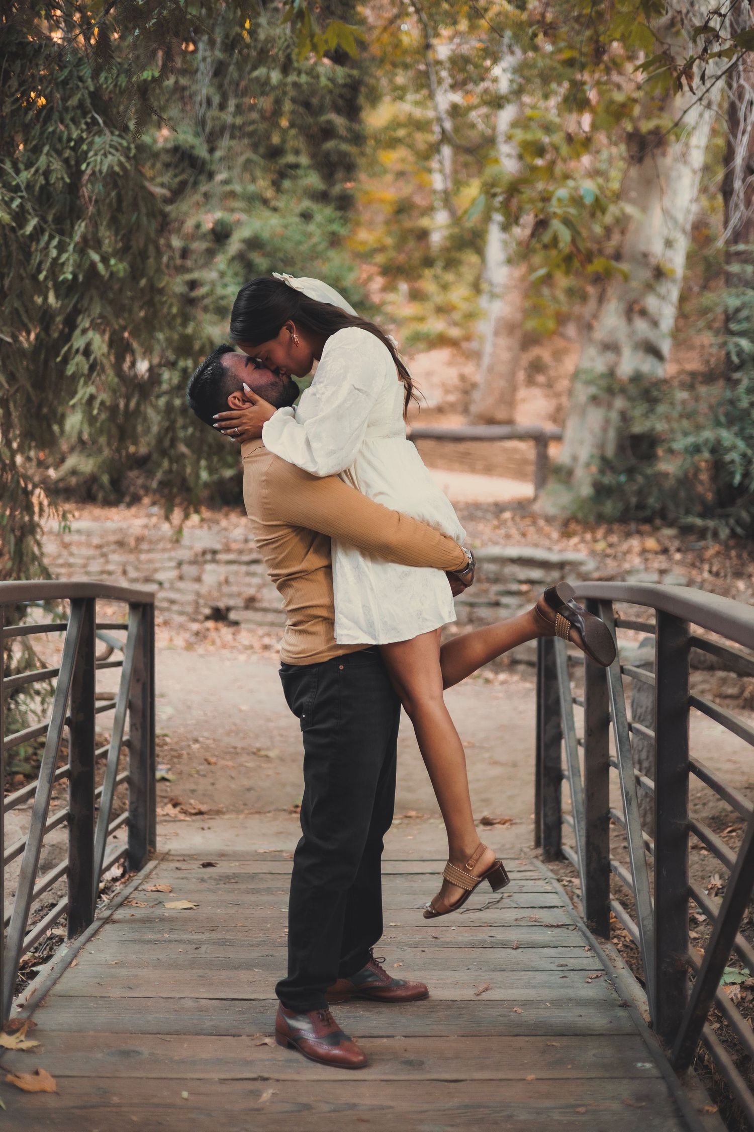 Romantic couple embrace and kiss while standing on rustic wooden bridge in autumn park setting.