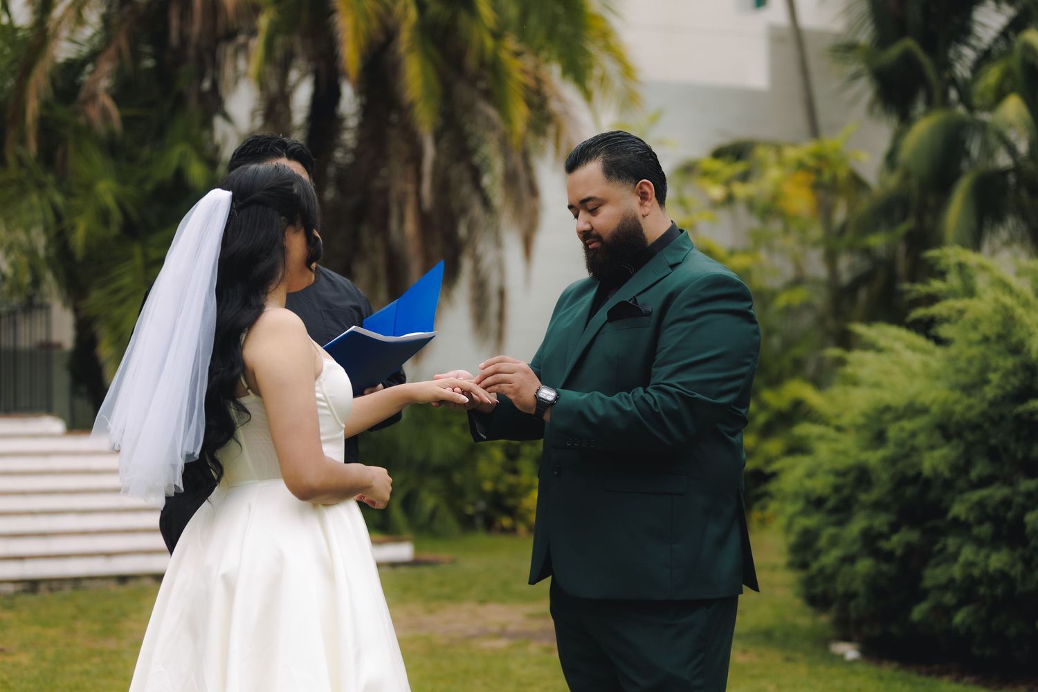 A couple in wedding attire shares a romantic moment in a tropical garden setting with palm trees.