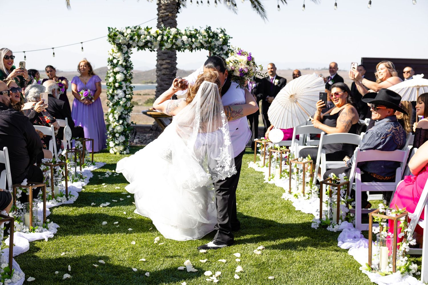 Wedding guests watch as a bride in a flowing white dress walks down an outdoor aisle lined with white chairs.