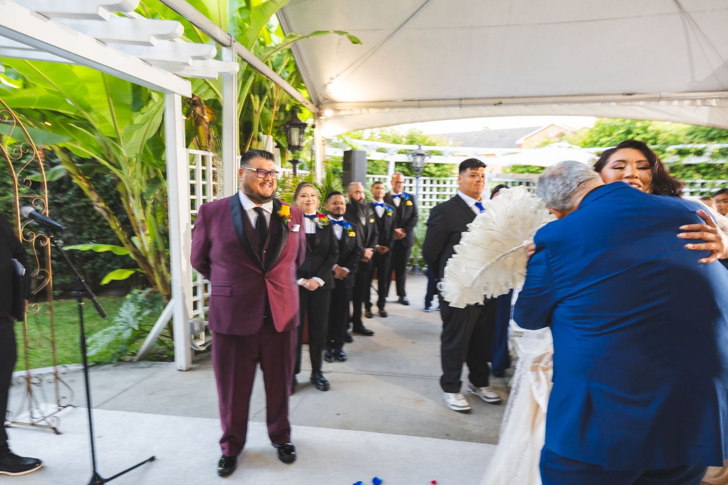 Wedding guests gather under an outdoor tent during a celebration with tropical greenery and decor.