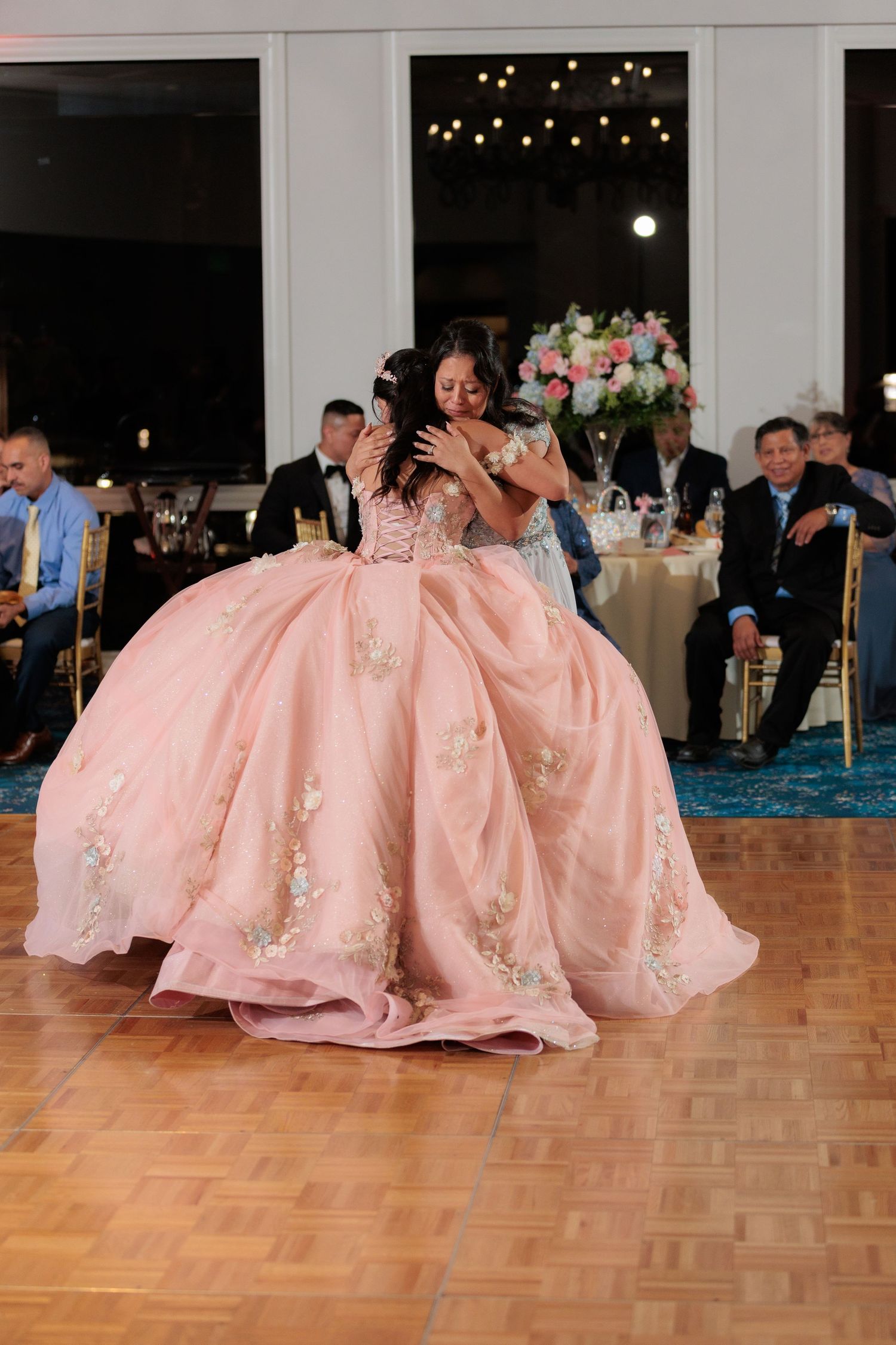 A group in pink formal dresses sits and laughs together on a wooden dance floor at an elegant reception venue.