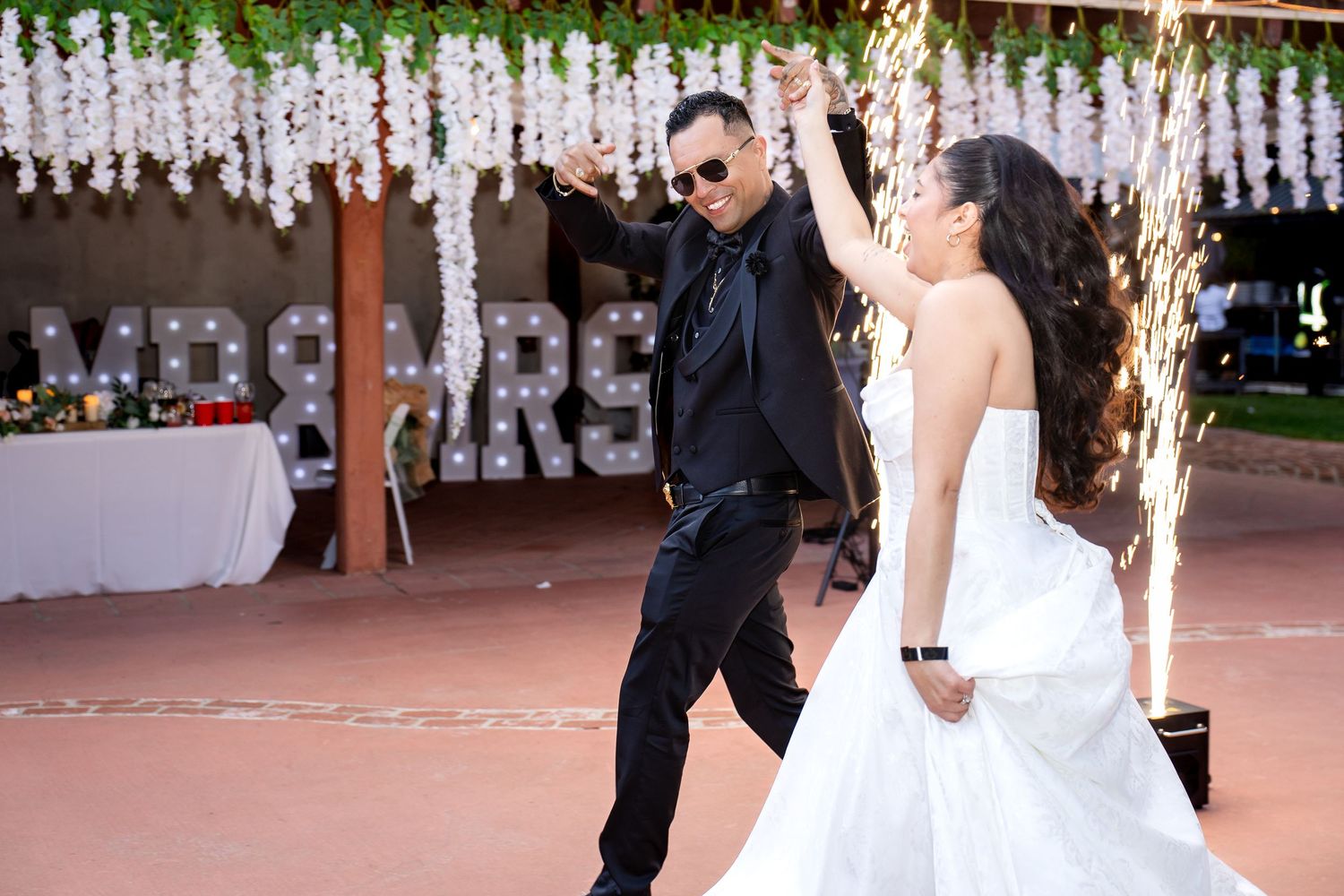 A couple dances together at a wedding reception under decorative hanging lights and floral arrangements.