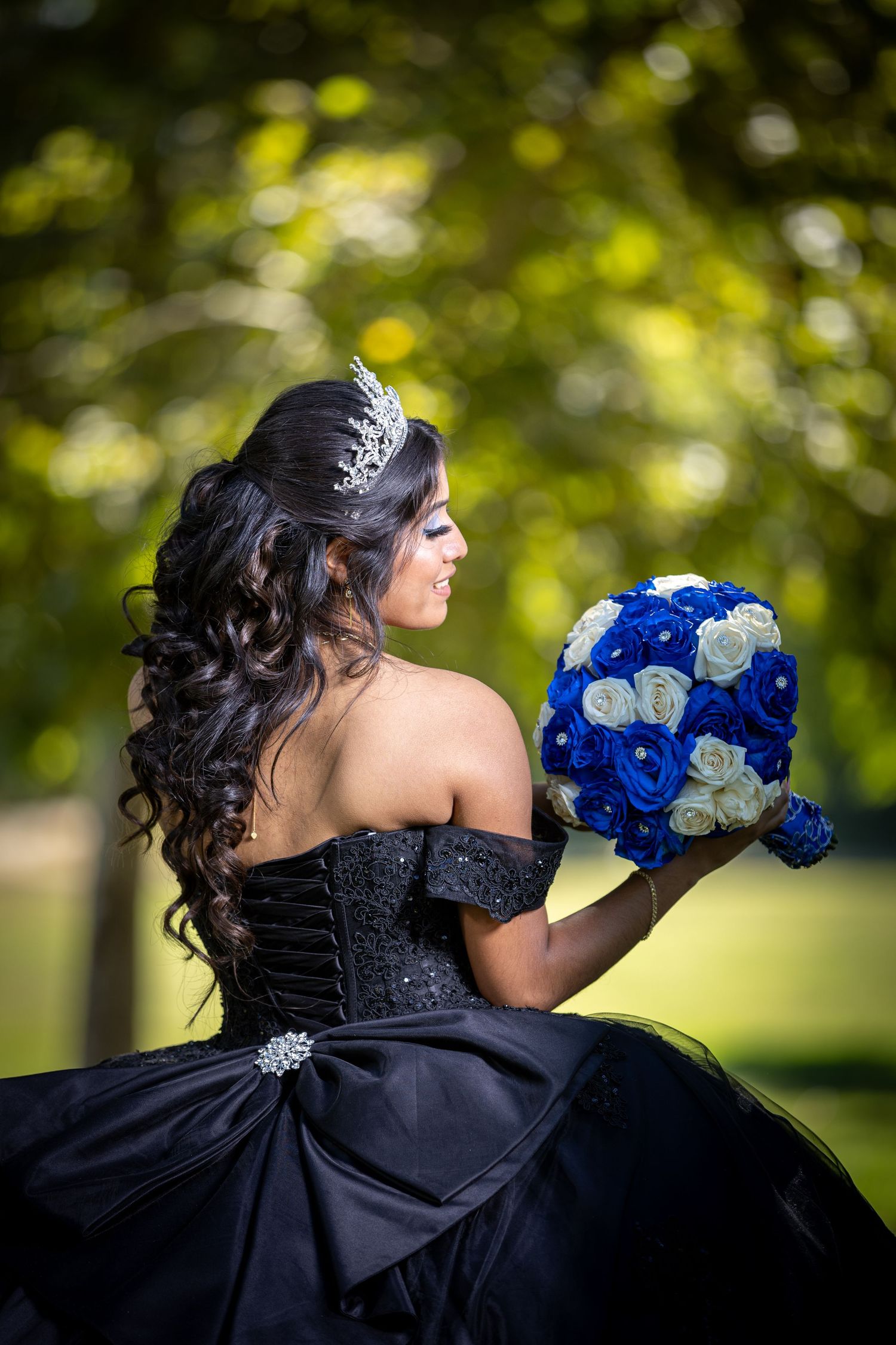 A bride in a black wedding dress holds a vibrant blue and white bouquet against a soft green outdoor background.