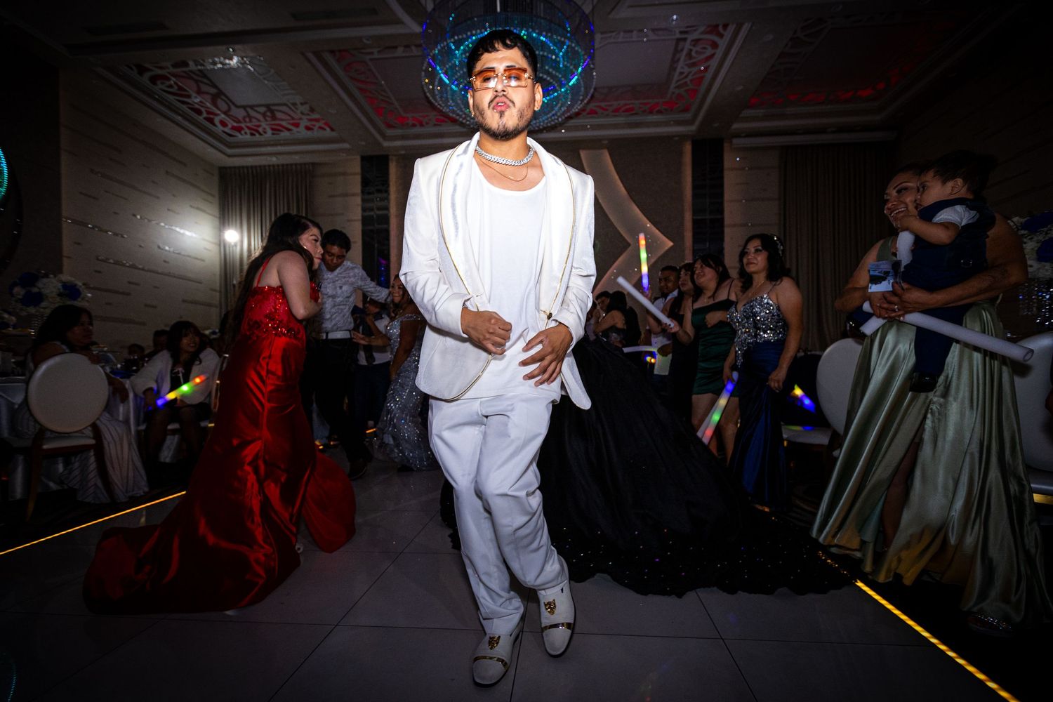 A person in white clothing performs a dance move in a dimly lit room with spectators watching from the sides.