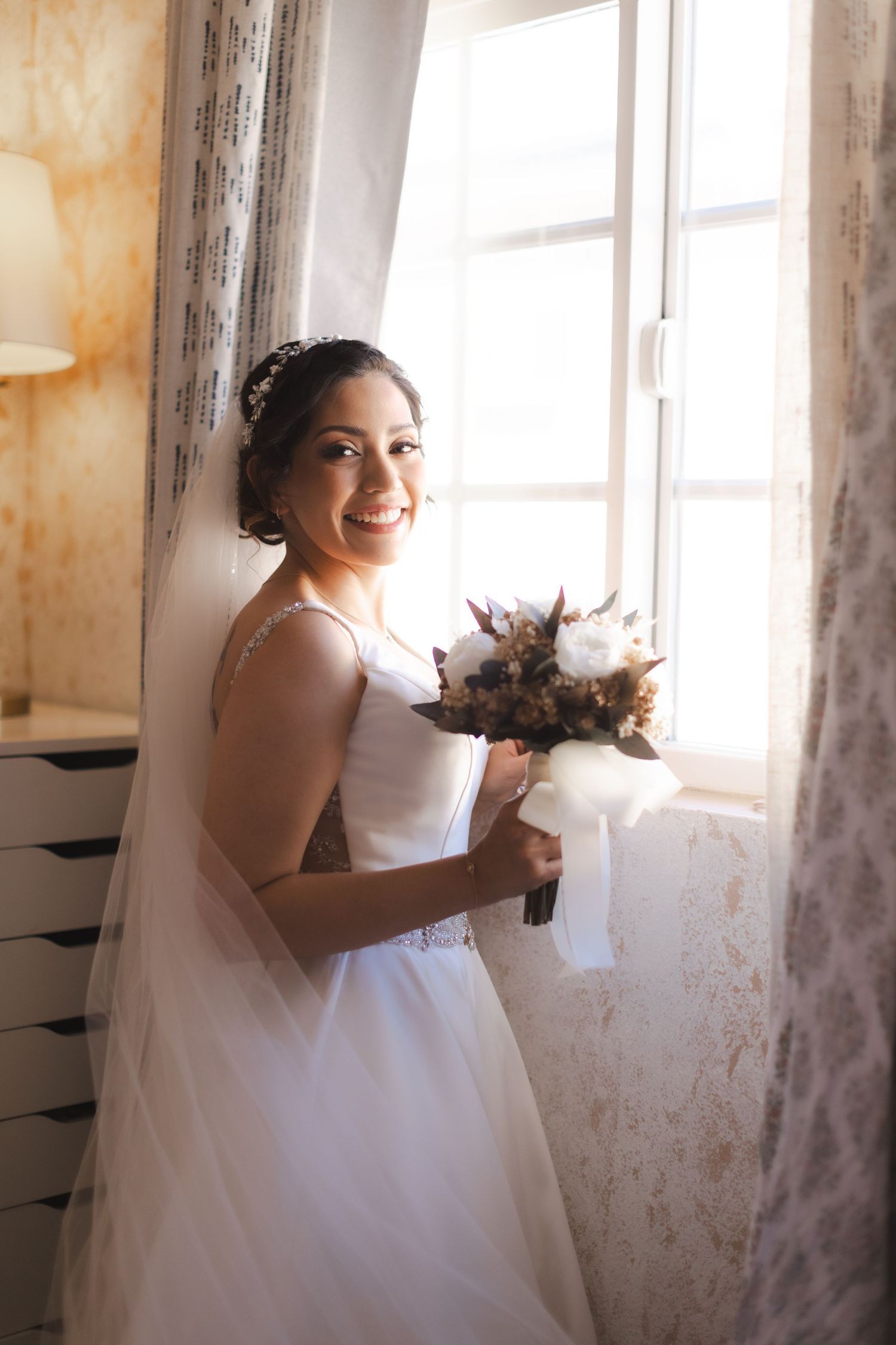 A bride in a white wedding dress smiles joyfully while holding her bouquet in a sunlit room.