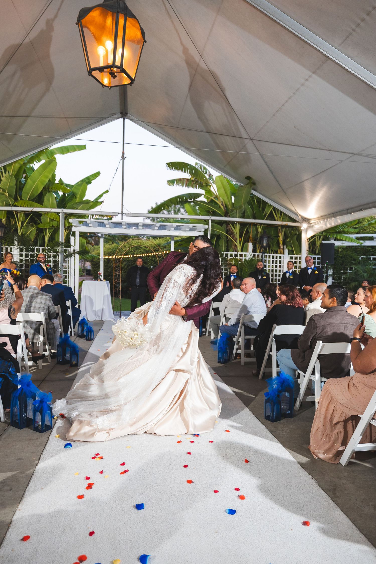 White wedding dress train decorated with red rose petals creates an elegant aisle runner under a covered outdoor venue.