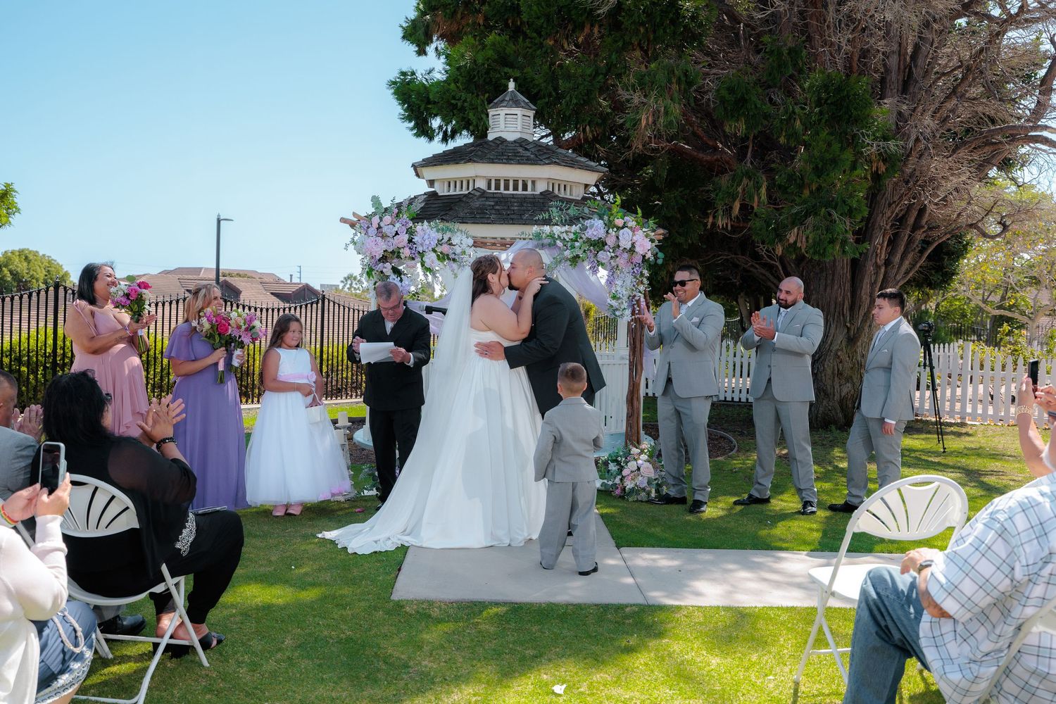 An outdoor spring wedding ceremony takes place under a white gazebo with guests seated in chairs on the lawn.