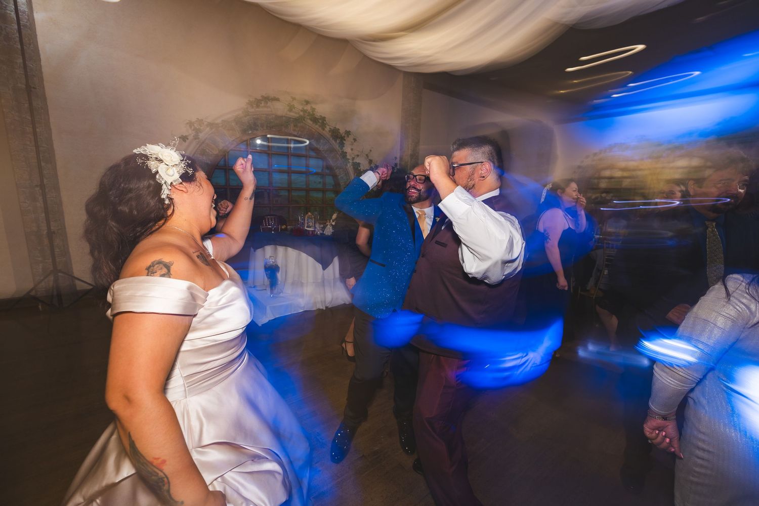 Wedding guests dance and celebrate on a dimly lit dance floor illuminated by blue lighting effects.