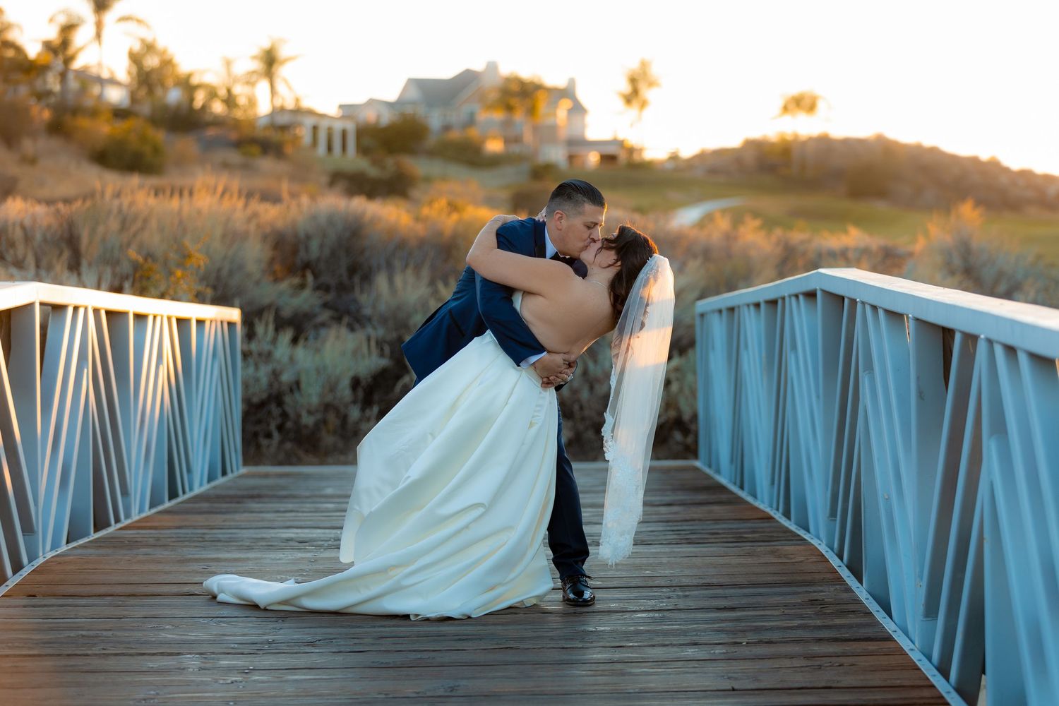 A romantic couple in wedding attire share a moment while dipping on a blue metal bridge at sunset.