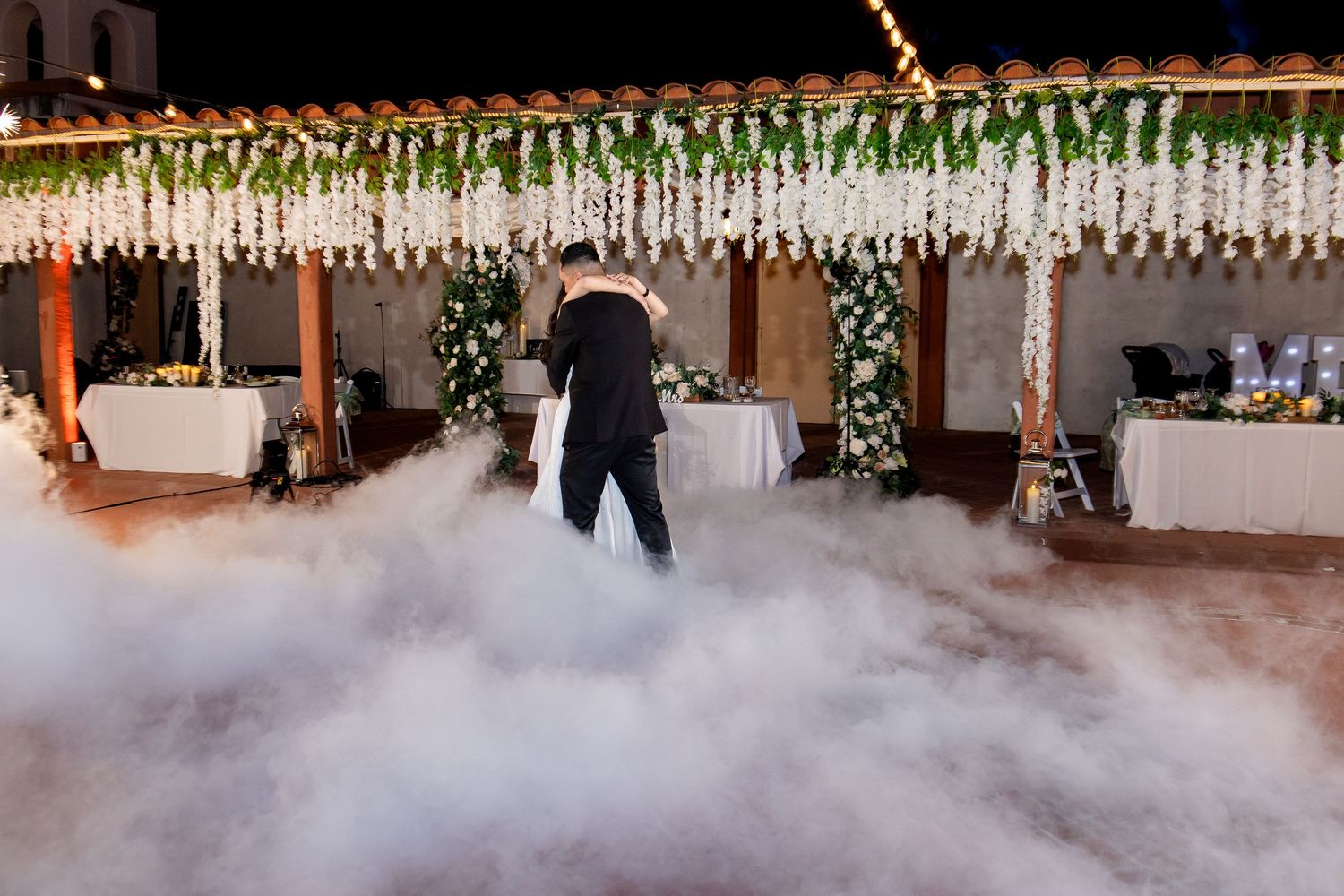 A wedding reception venue decorated with elegant white floral arrangements and ambient fog effects on the dance floor.