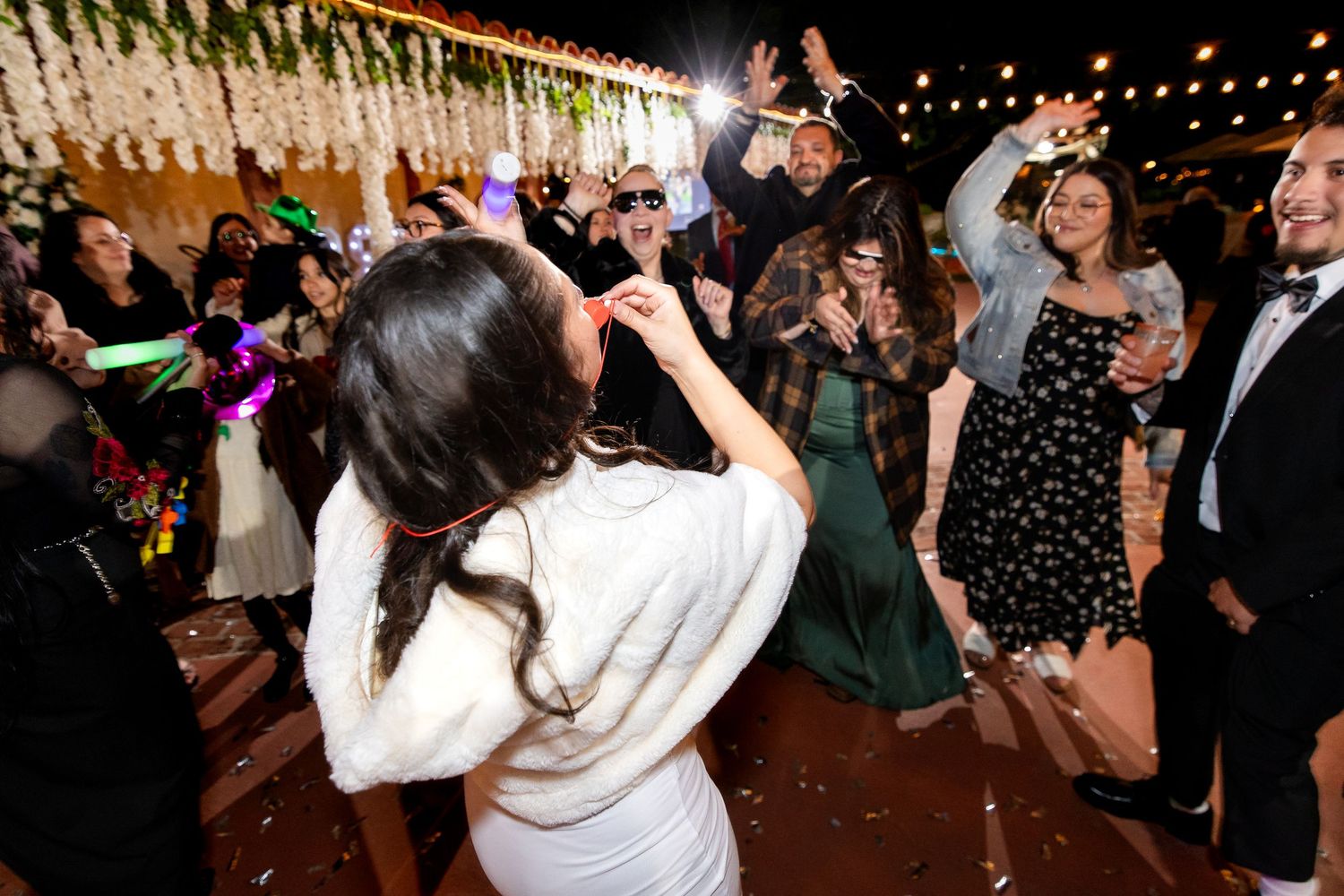 People dancing and celebrating at a lively wedding reception under string lights.