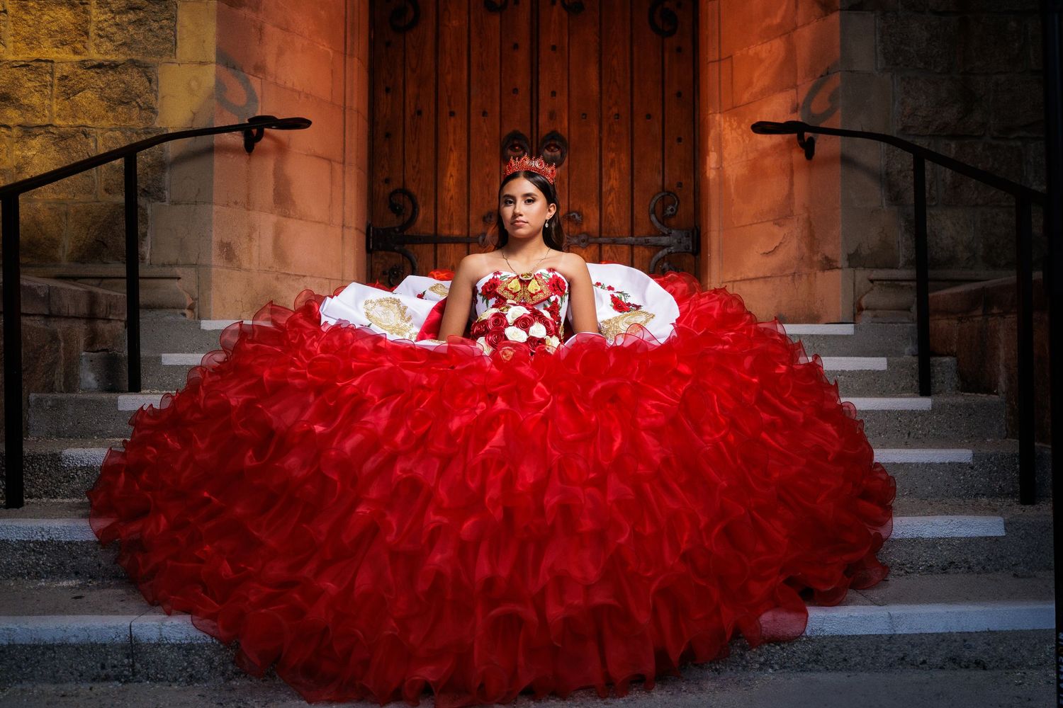 A red tulle ballgown dress with layers of ruffles is photographed against stone architecture in an outdoor setting.
