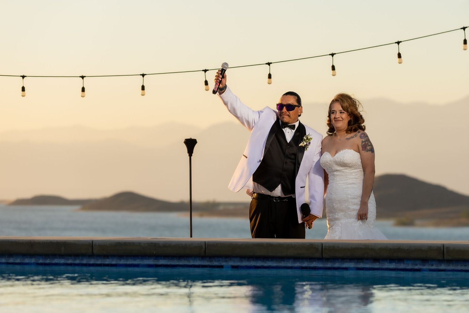 Couple celebrates at sunset by a pool with string lights and mountain views in the background.