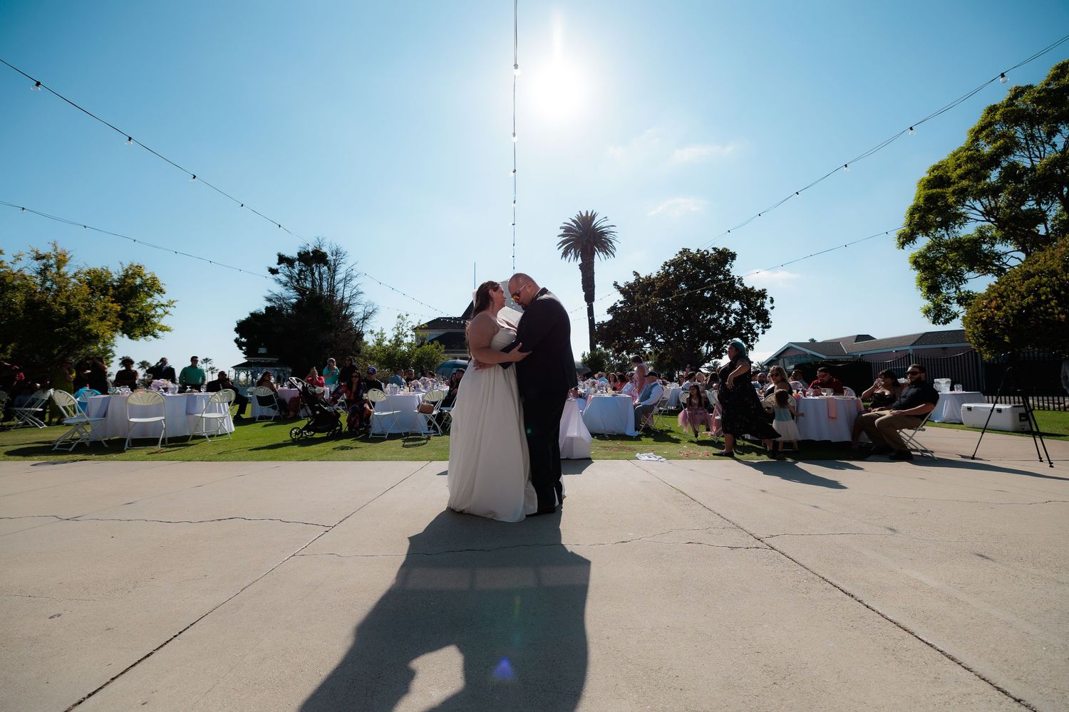 A couple shares a romantic moment during their outdoor wedding ceremony under bright sunlight on a paved courtyard.