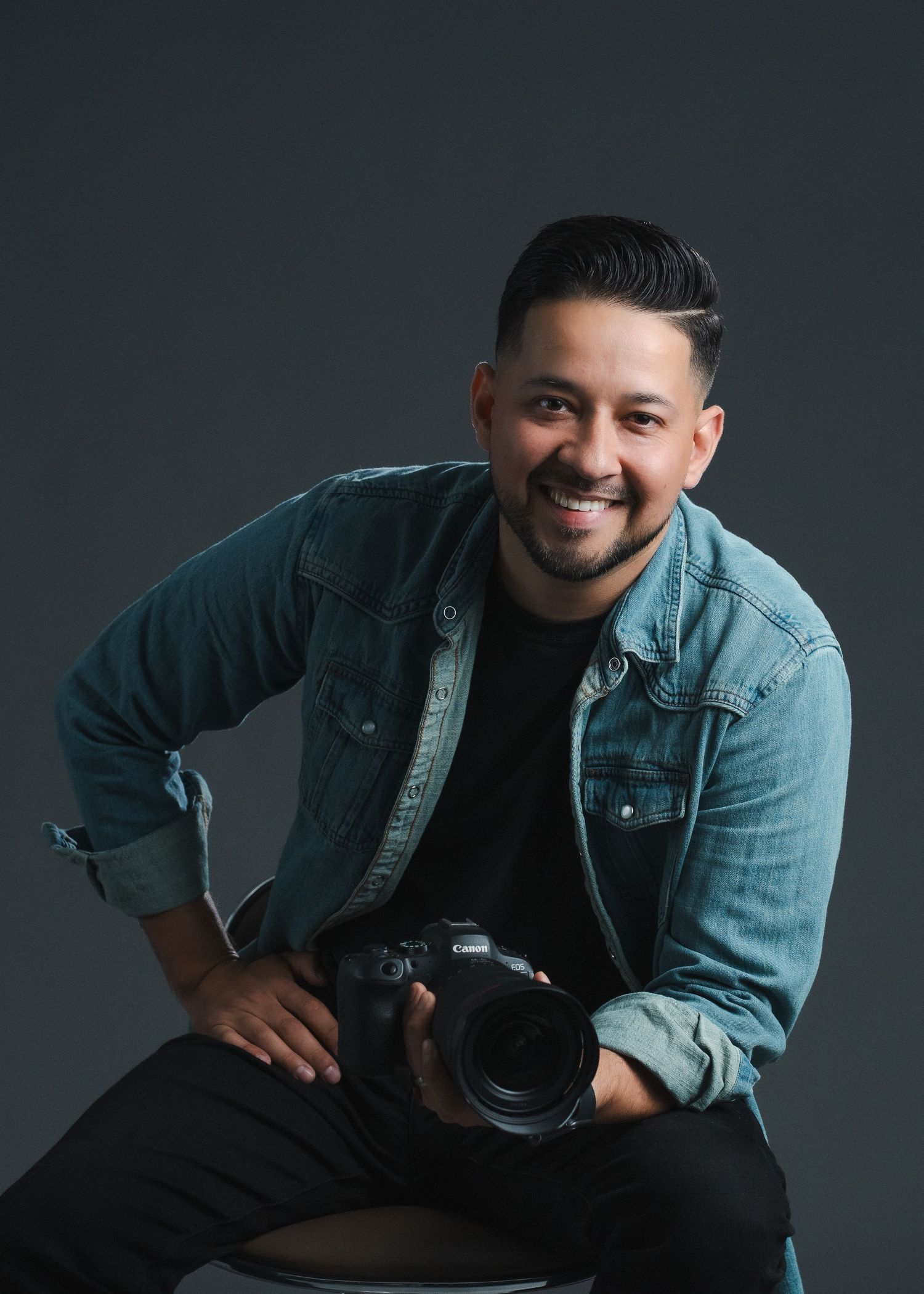 Professional photographer in denim jacket poses with camera against dark background.