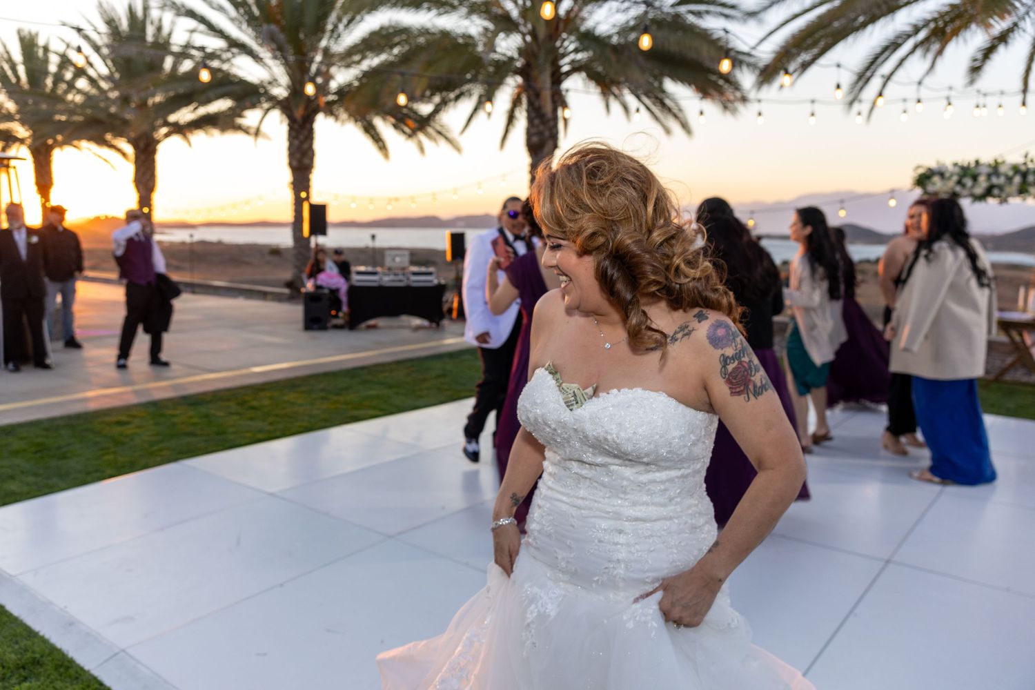 A group of people dancing and celebrating at an outdoor wedding reception during sunset near palm trees.