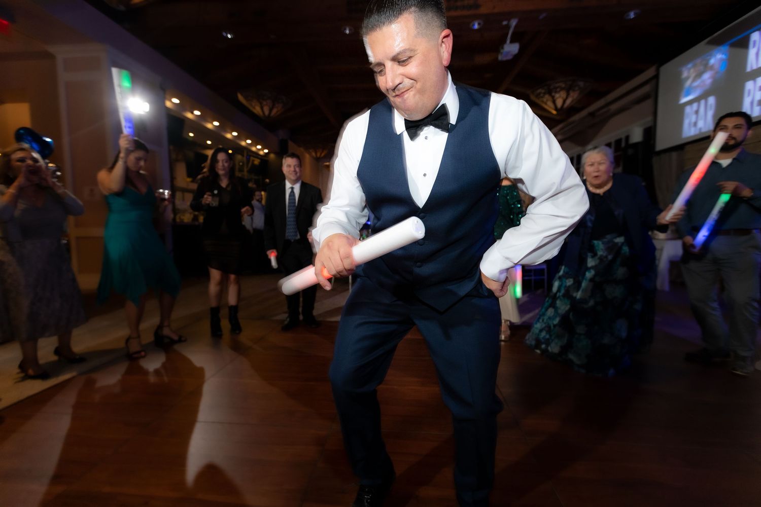 Someone in a dress shirt and vest dances energetically on a dimly lit dance floor during a formal event.