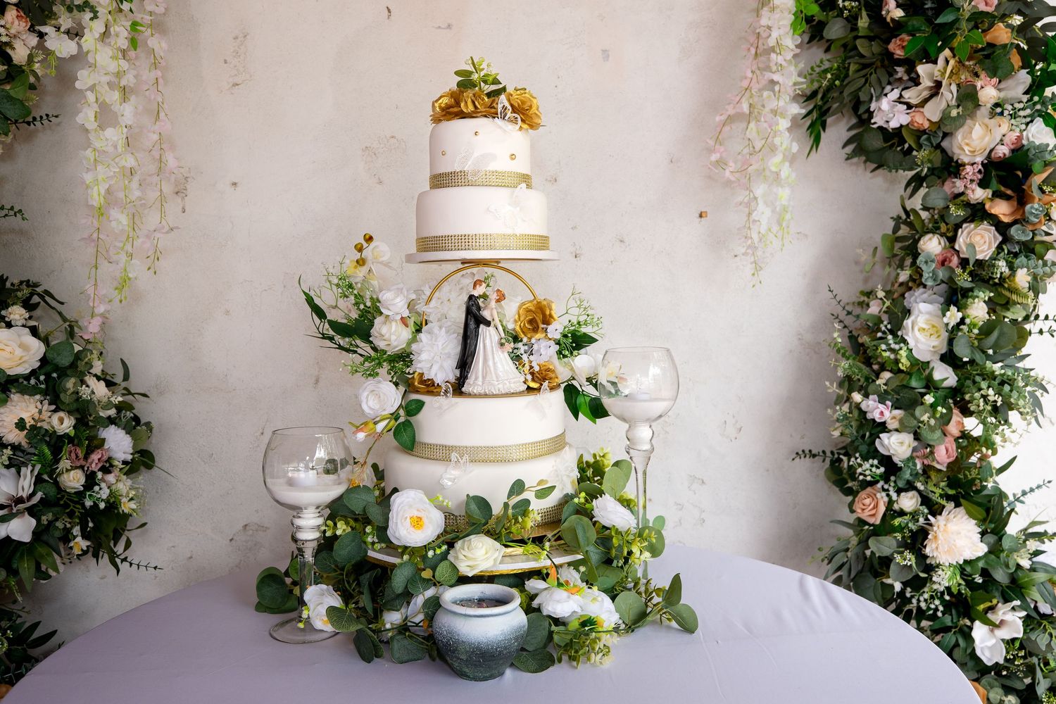 Elegant three-tier white wedding cake decorated with fresh flowers and greenery on display table with floral arrangements.