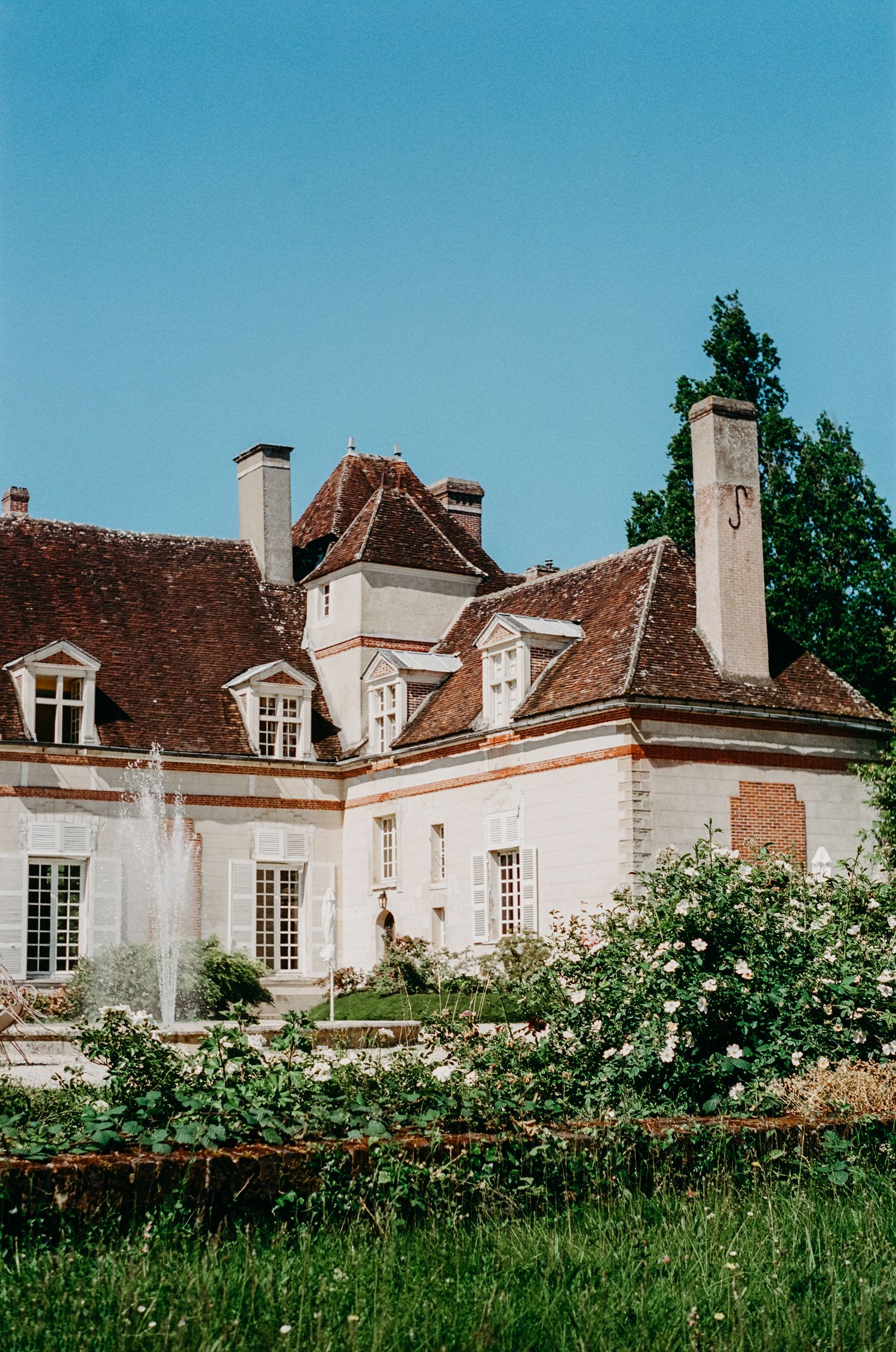 French country manor house with terracotta roof and dormer windows surrounded by blooming garden.