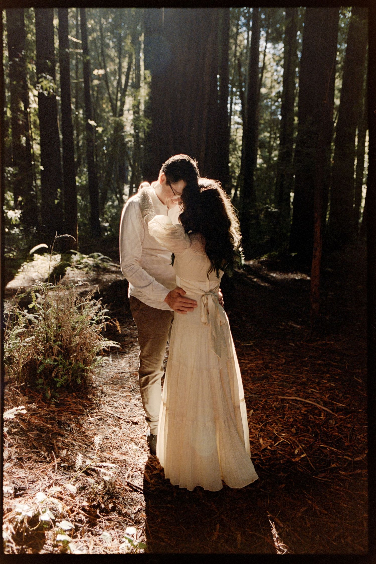 A couple shares a romantic kiss in a sunlit forest, the bride wearing an elegant white gown.