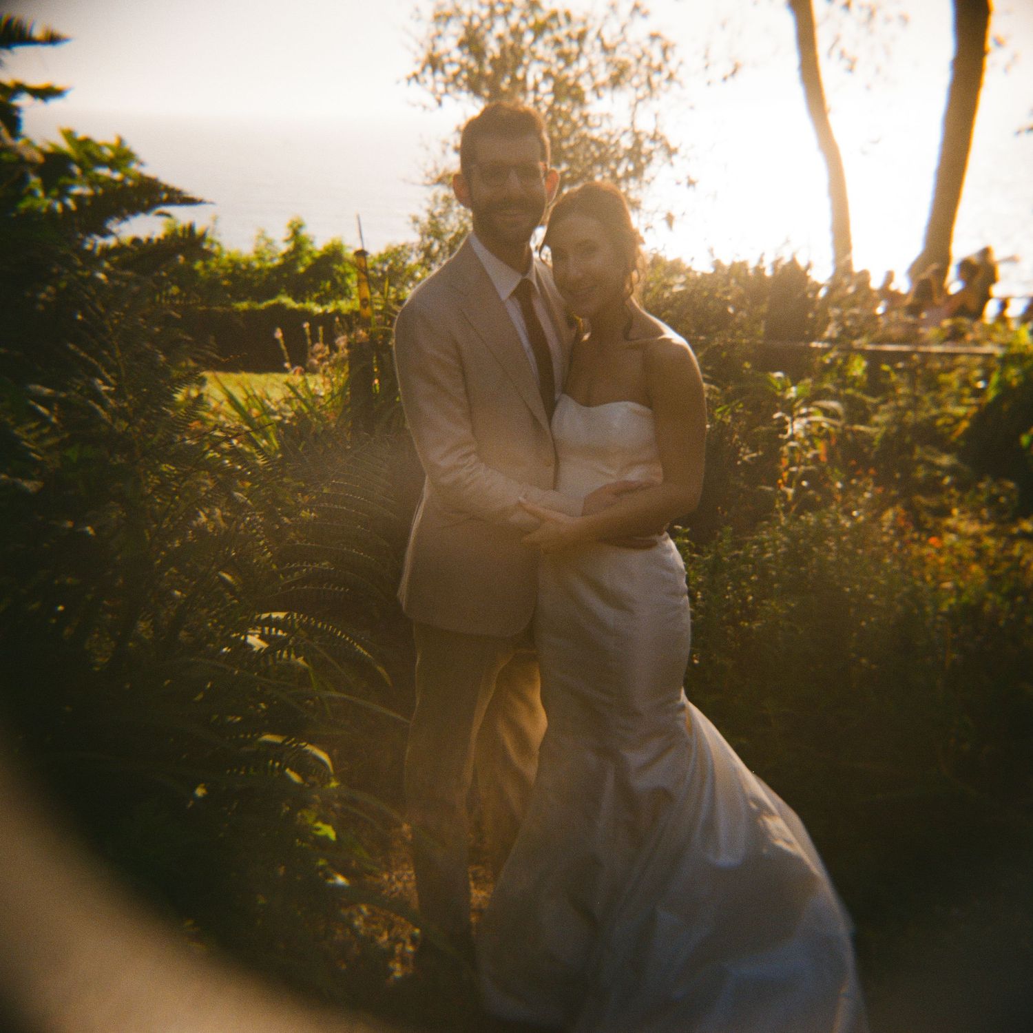 A couple in wedding attire embraces in a lush garden at golden hour, the bride in white gown and groom in tan suit.