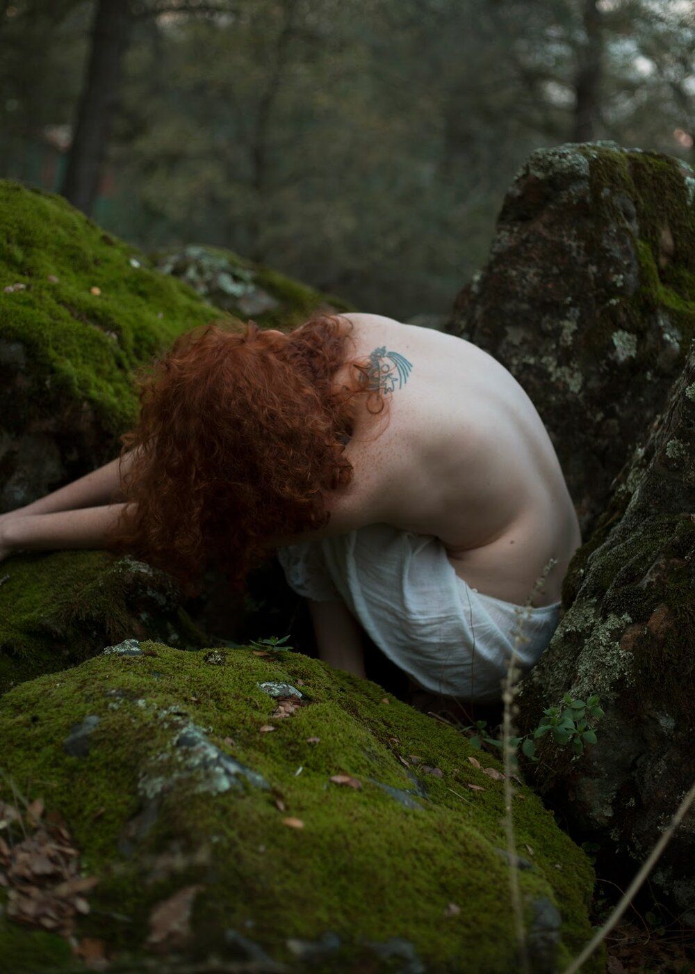 Artistic photo of figure in white dress resting against moss-covered rocks in forest setting.