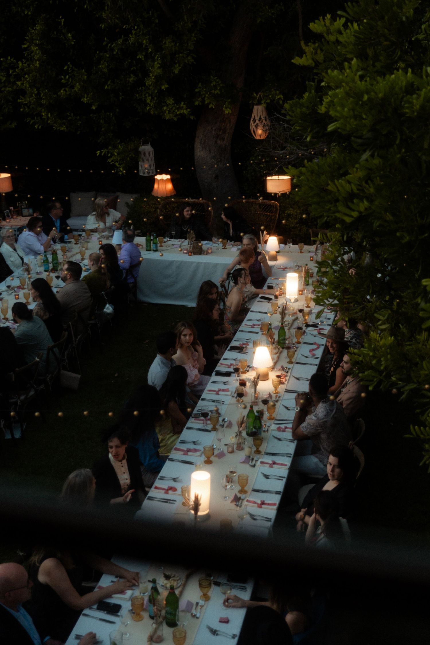 An elegant outdoor dinner setting with long tables illuminated by candlelight and lanterns.