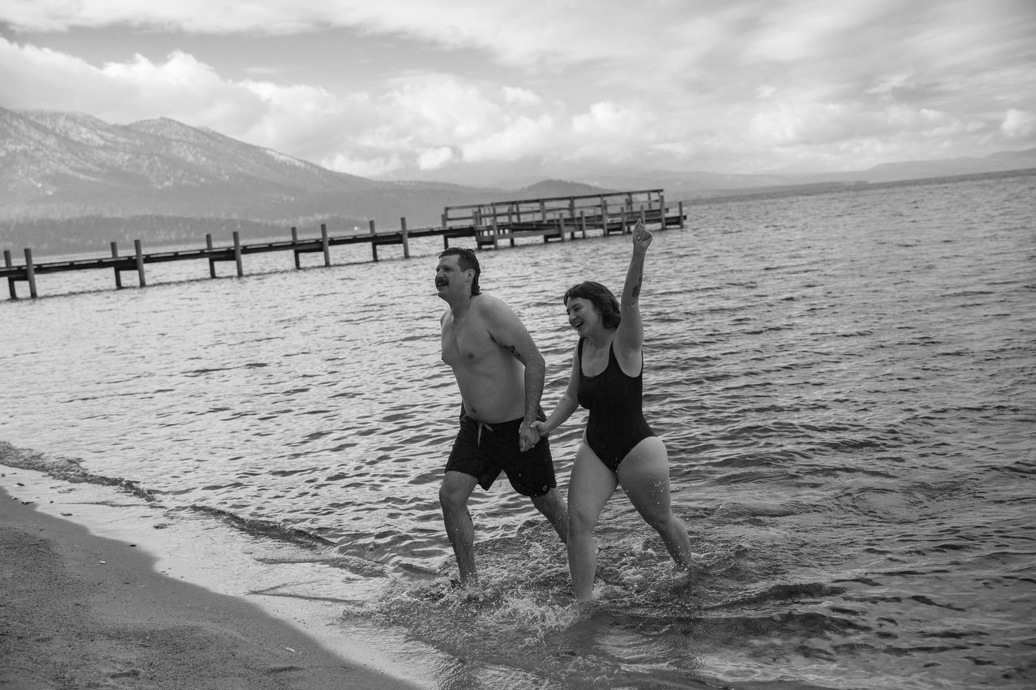 A playful couple splashing in the ocean water near a long wooden pier.