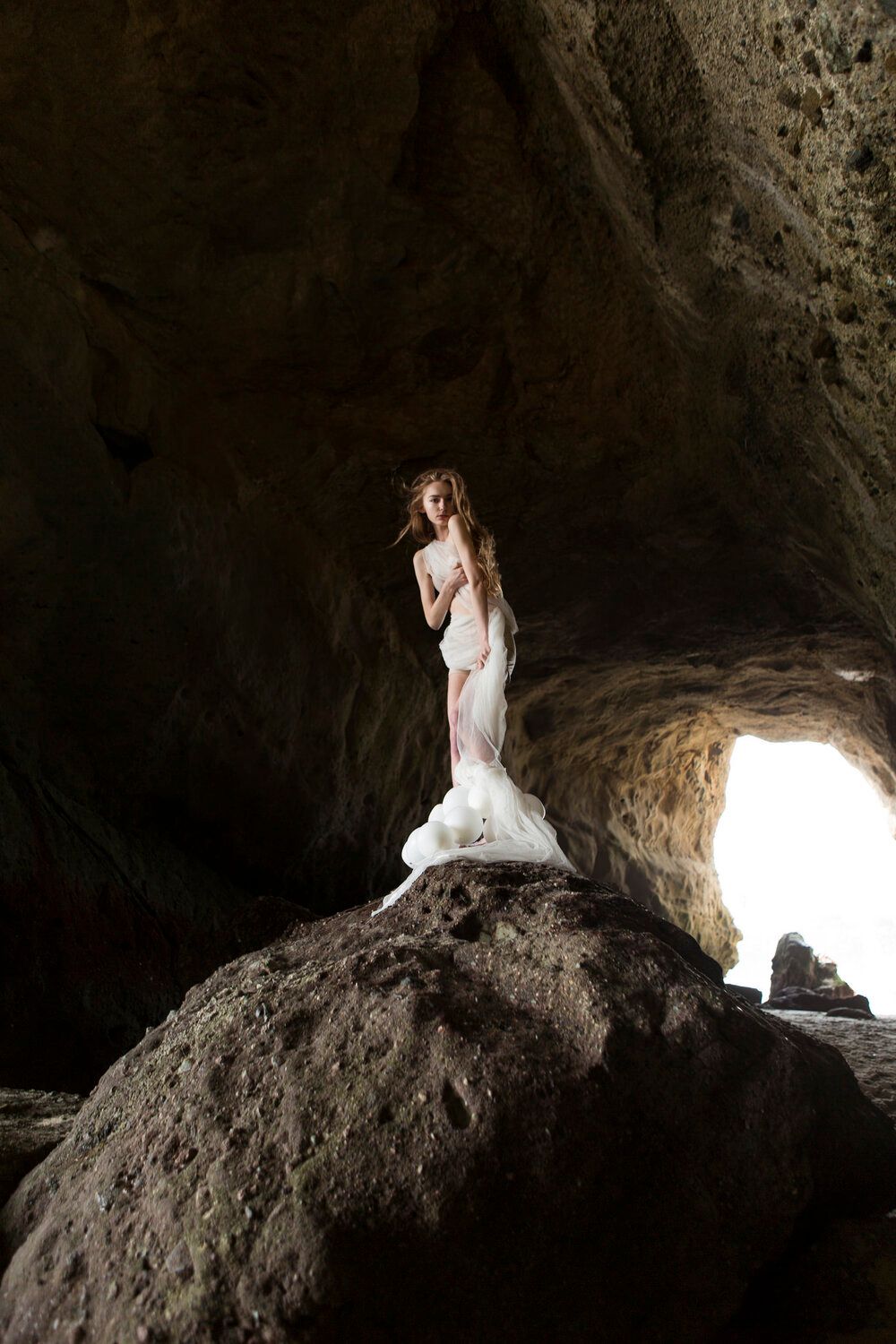 A figure in white dress sits dramatically posed in a dark cave opening with natural light streaming in.