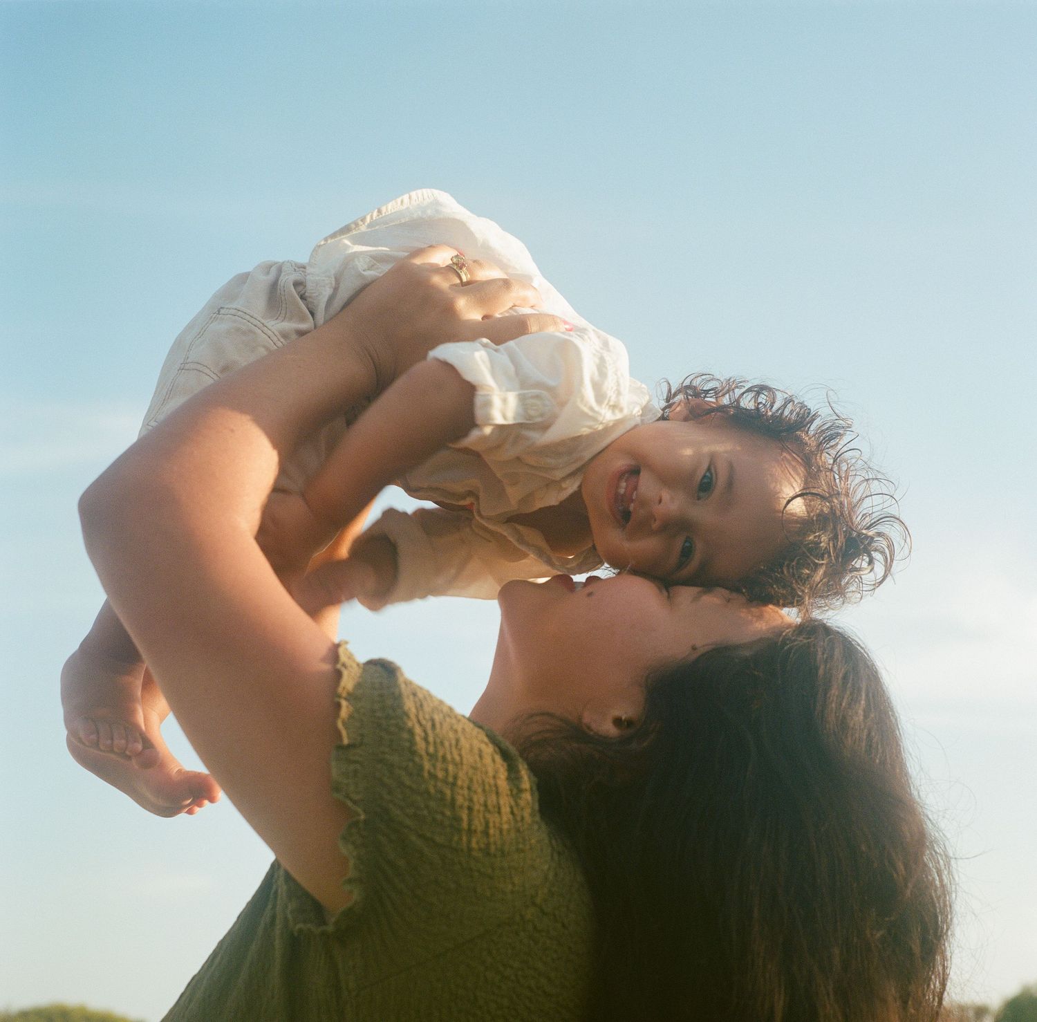 A joyful mother lifts her laughing baby girl overhead against a bright blue sky outdoors.