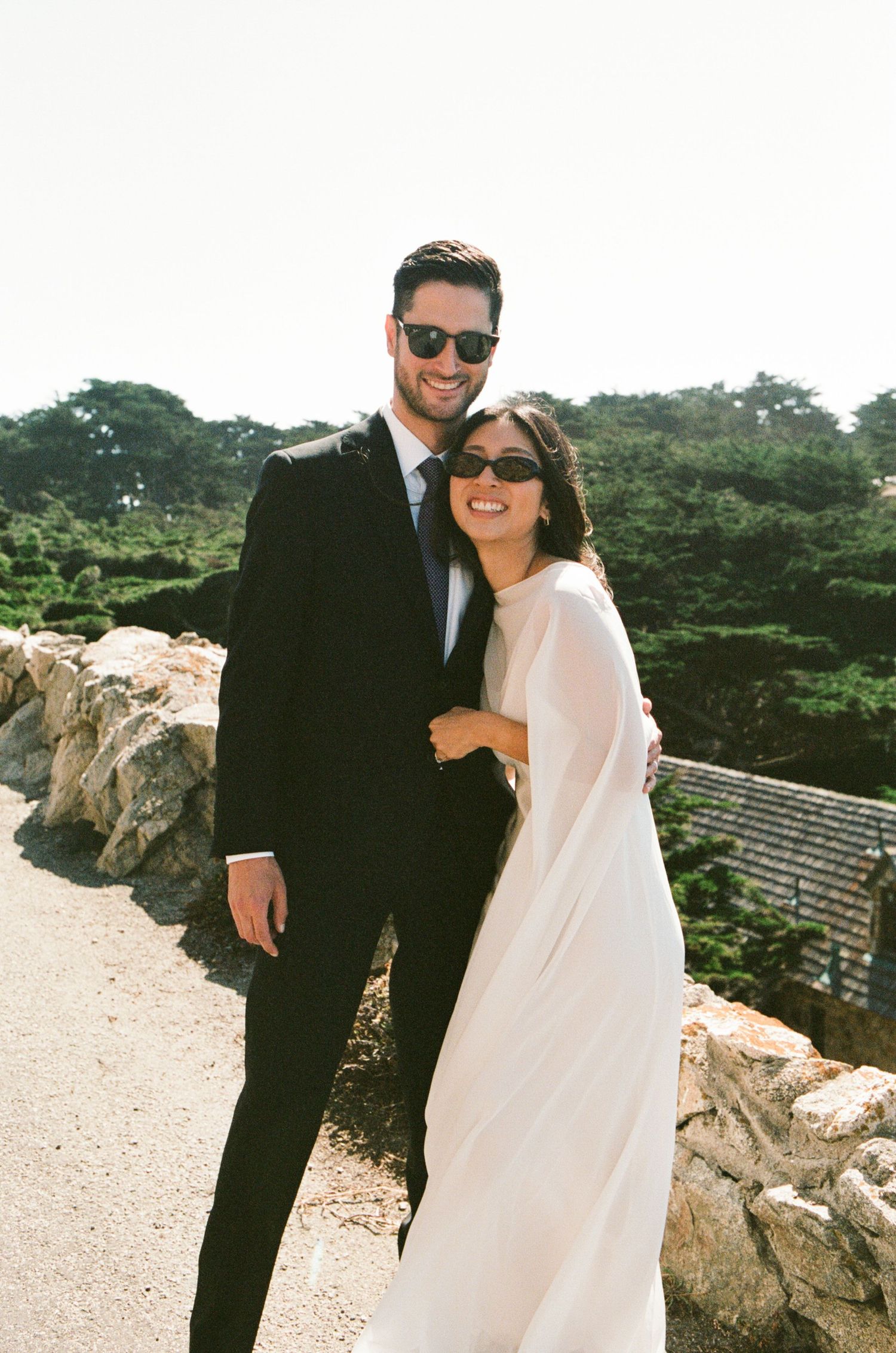 A couple in wedding attire with sunglasses pose outdoors near stone walls and lush trees on their wedding day.