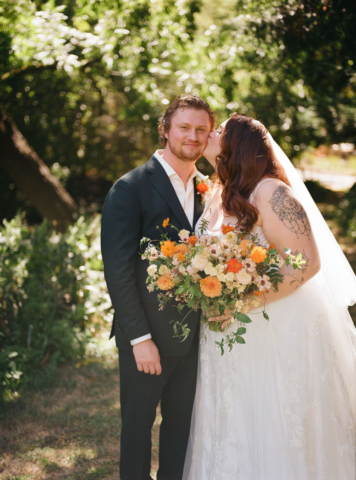 A romantic outdoor wedding scene with a couple sharing a moment among greenery and holding a peach and white bouquet.