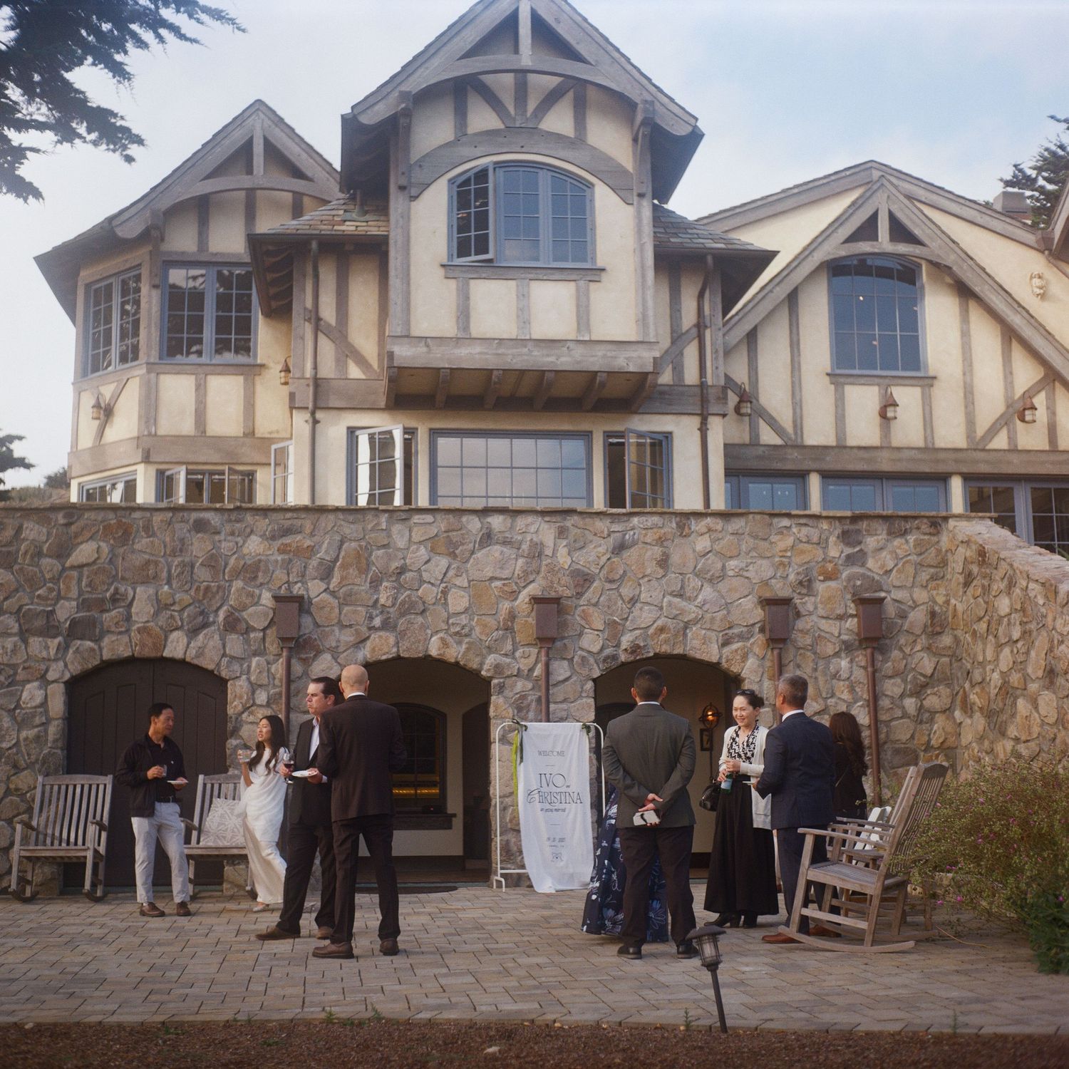 People socializing outside a large Tudor-style stone and timber building during an outdoor gathering event.
