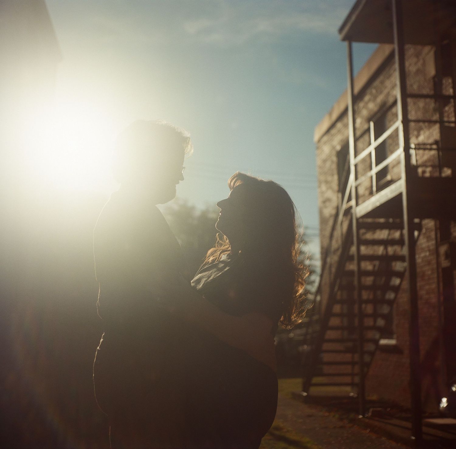 Couple embracing in golden sunlight outdoors near stone building with metal staircase on a clear blue sky day.