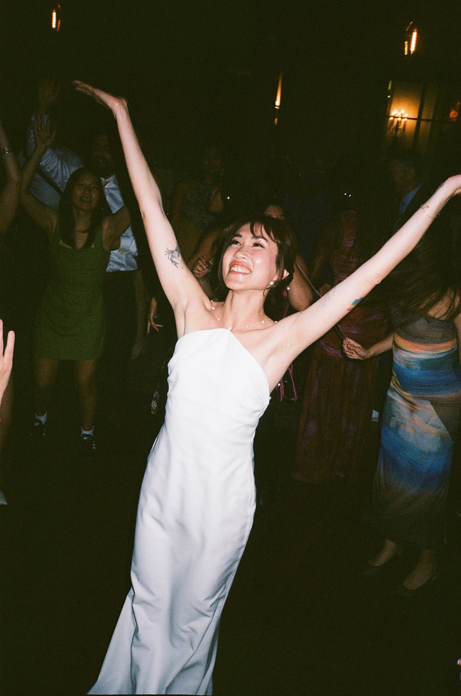 Person in white dress dances joyfully with arms raised at a dimly lit nighttime party or event.